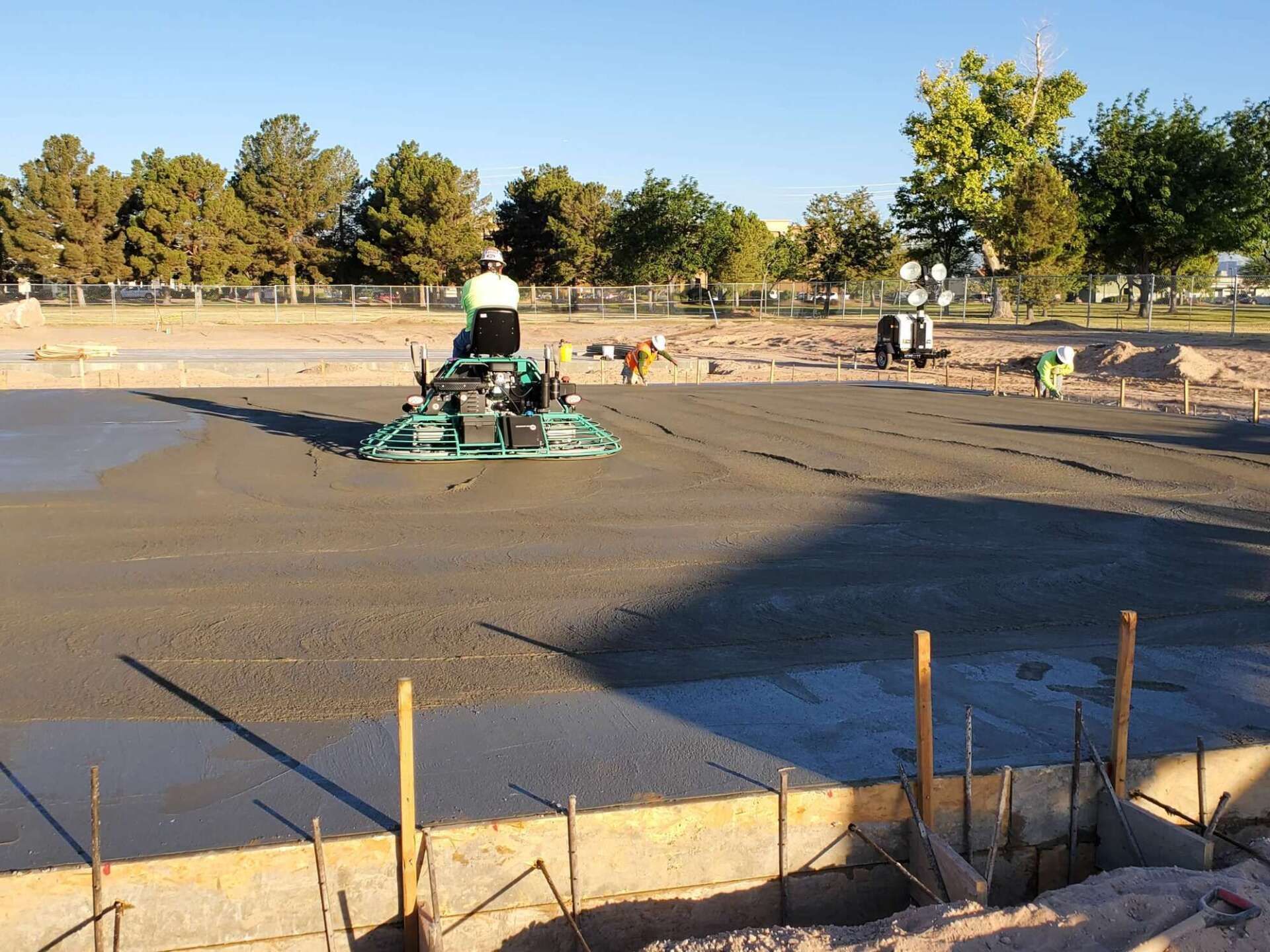 Construction worker smoothing wet concrete with a power trowel; outdoors, trees in background.