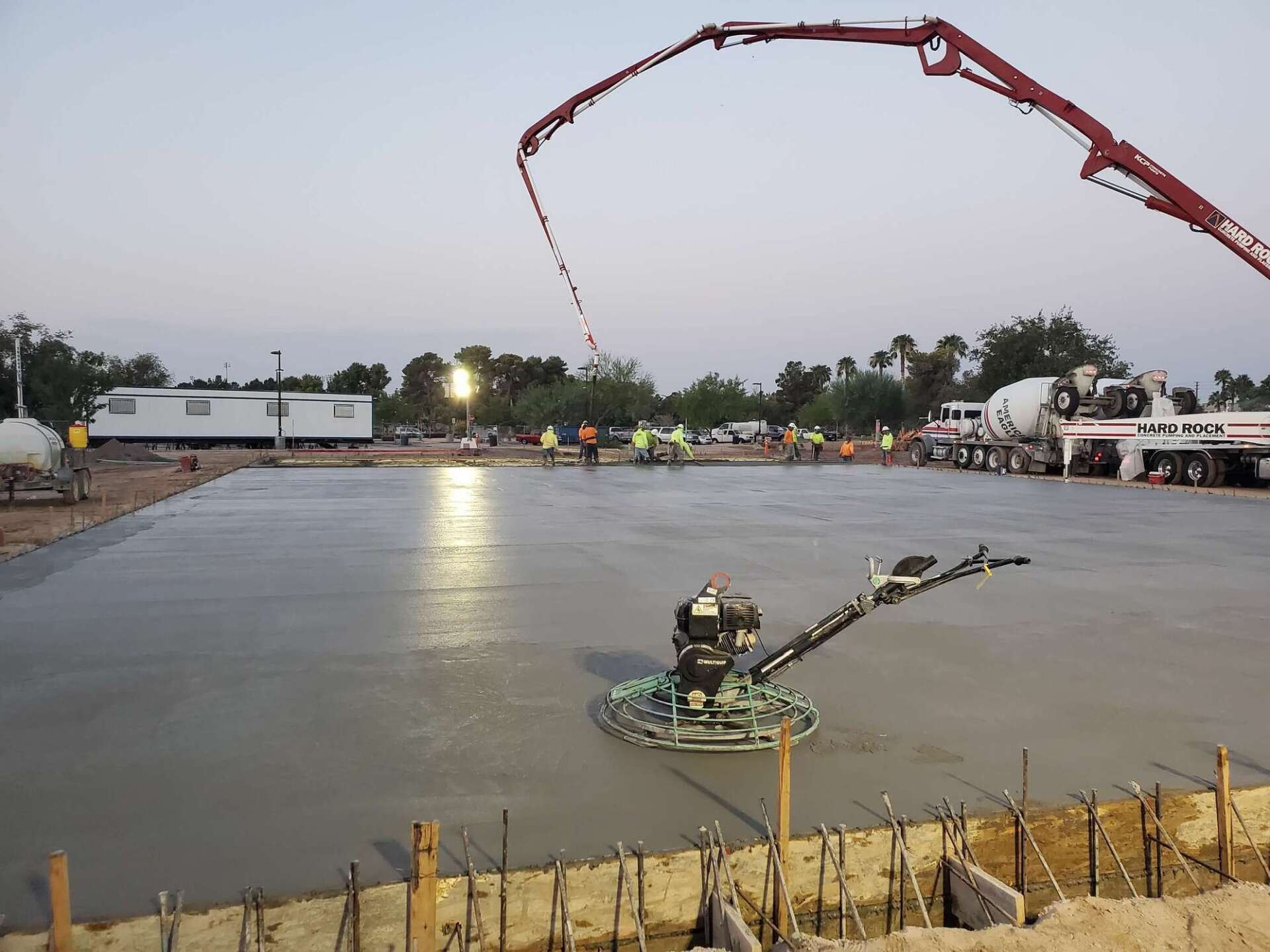 Concrete pour at dusk. A power trowel smooths the wet concrete. A pump truck and workers are present.