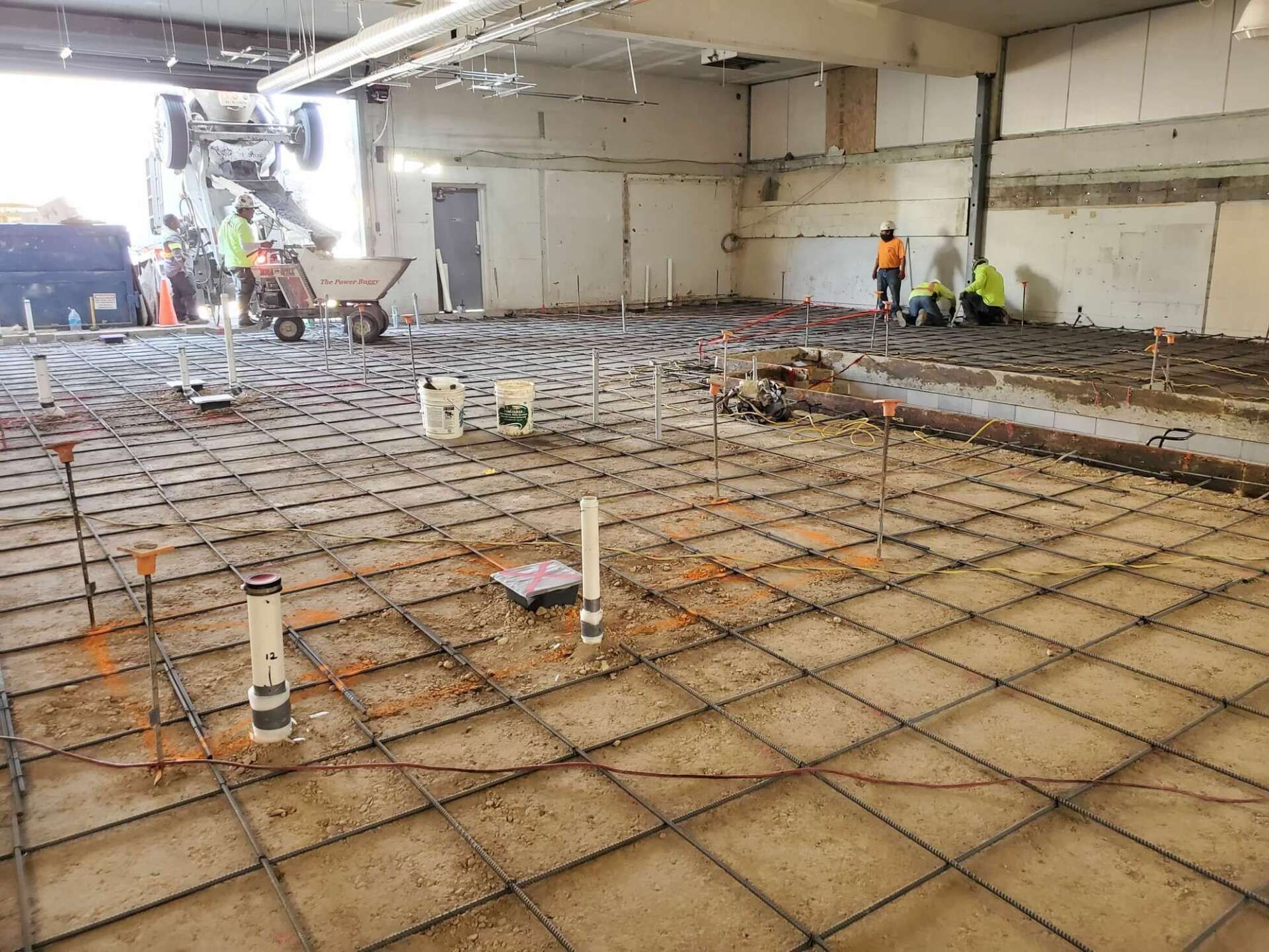 Construction site with workers, rebar grid, plumbing, and concrete being poured.