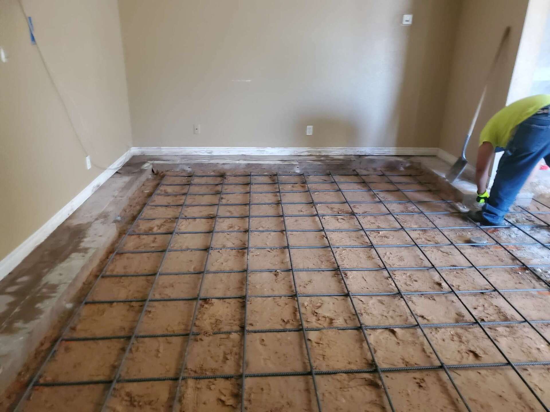 Construction worker installing rebar grid on a dirt floor, preparing for concrete. Room with beige walls.