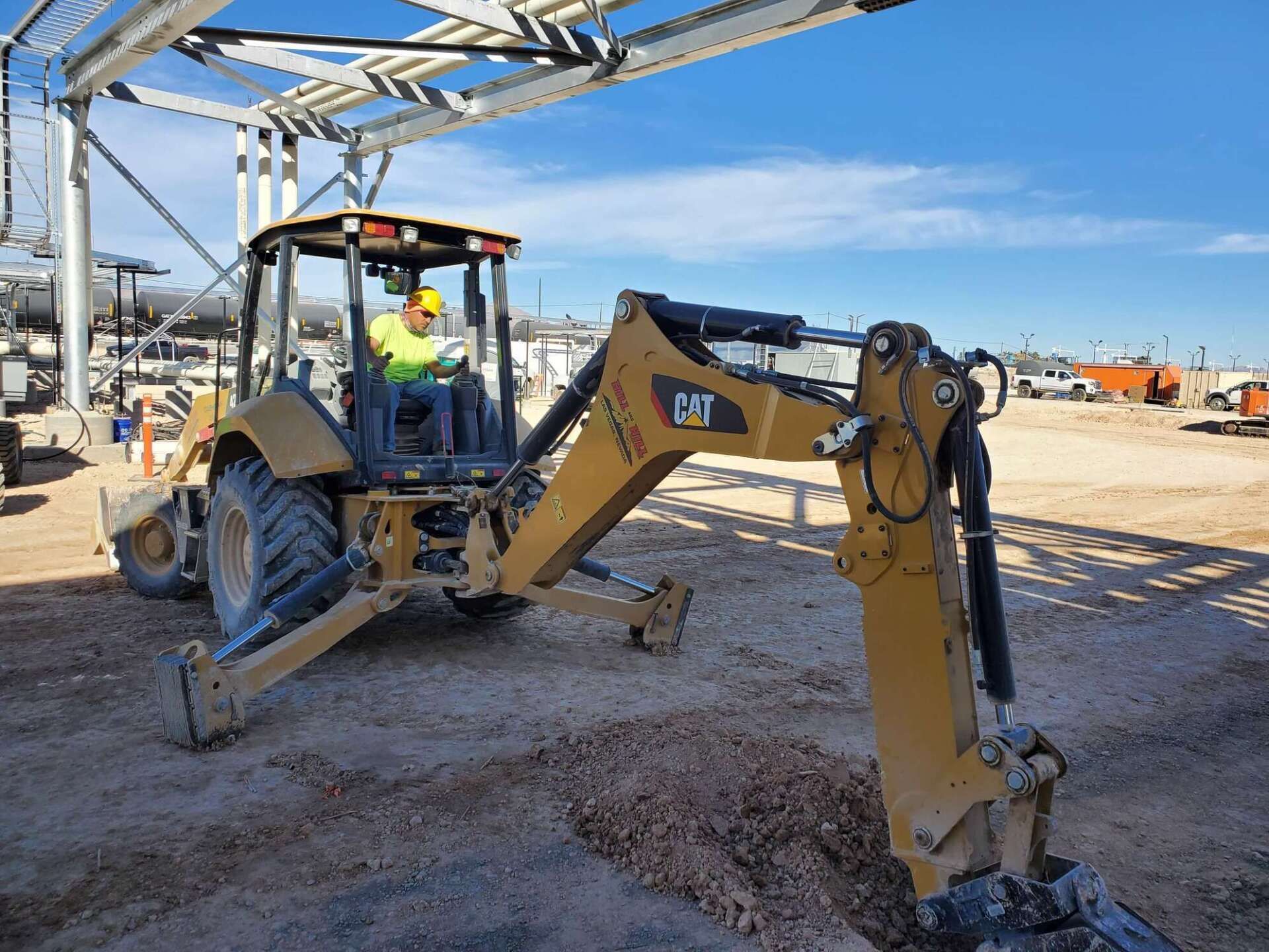 A yellow CAT backhoe digging in a construction site with a worker in the cab. Blue sky in background.