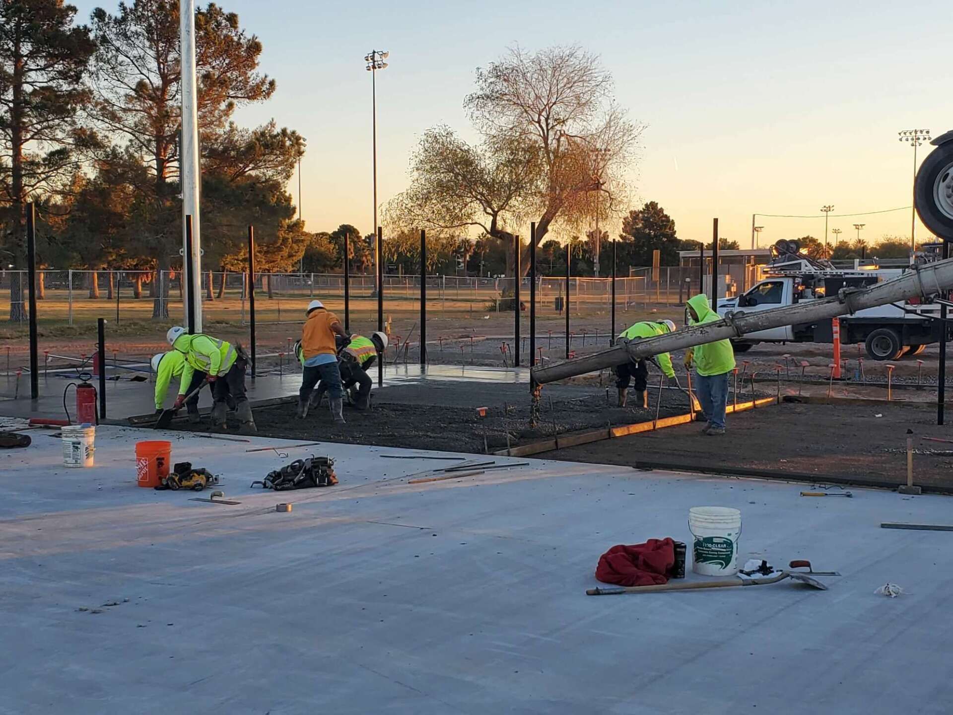 Construction workers pouring concrete at a park with fence posts and a truck. Setting sun in background.