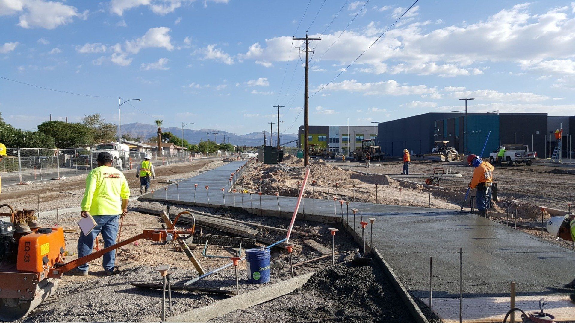 Construction site with workers pouring concrete; road and sidewalk work in progress.