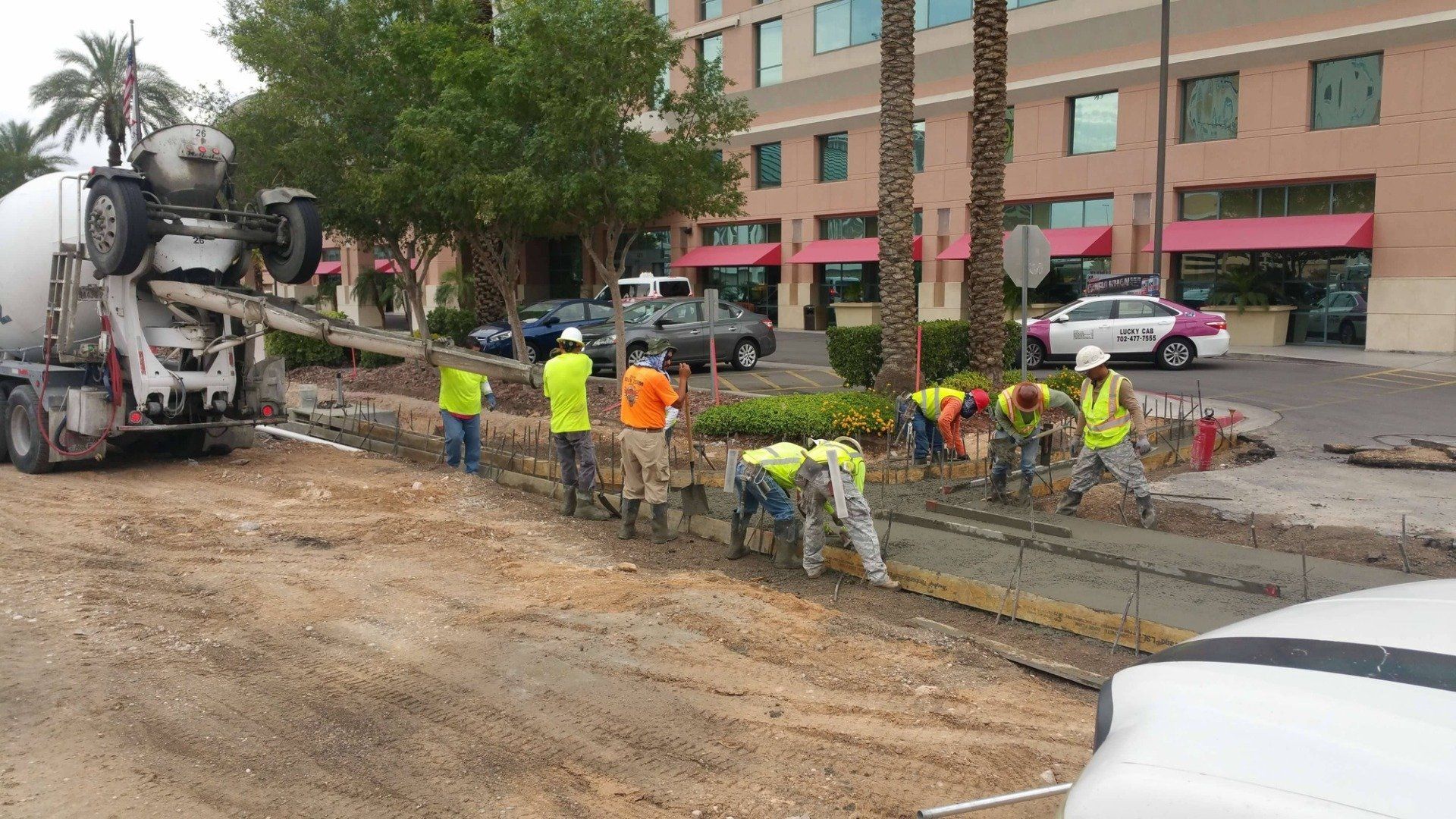 Construction workers pouring concrete on a sidewalk. Cement truck, people in safety vests, hotel in background.