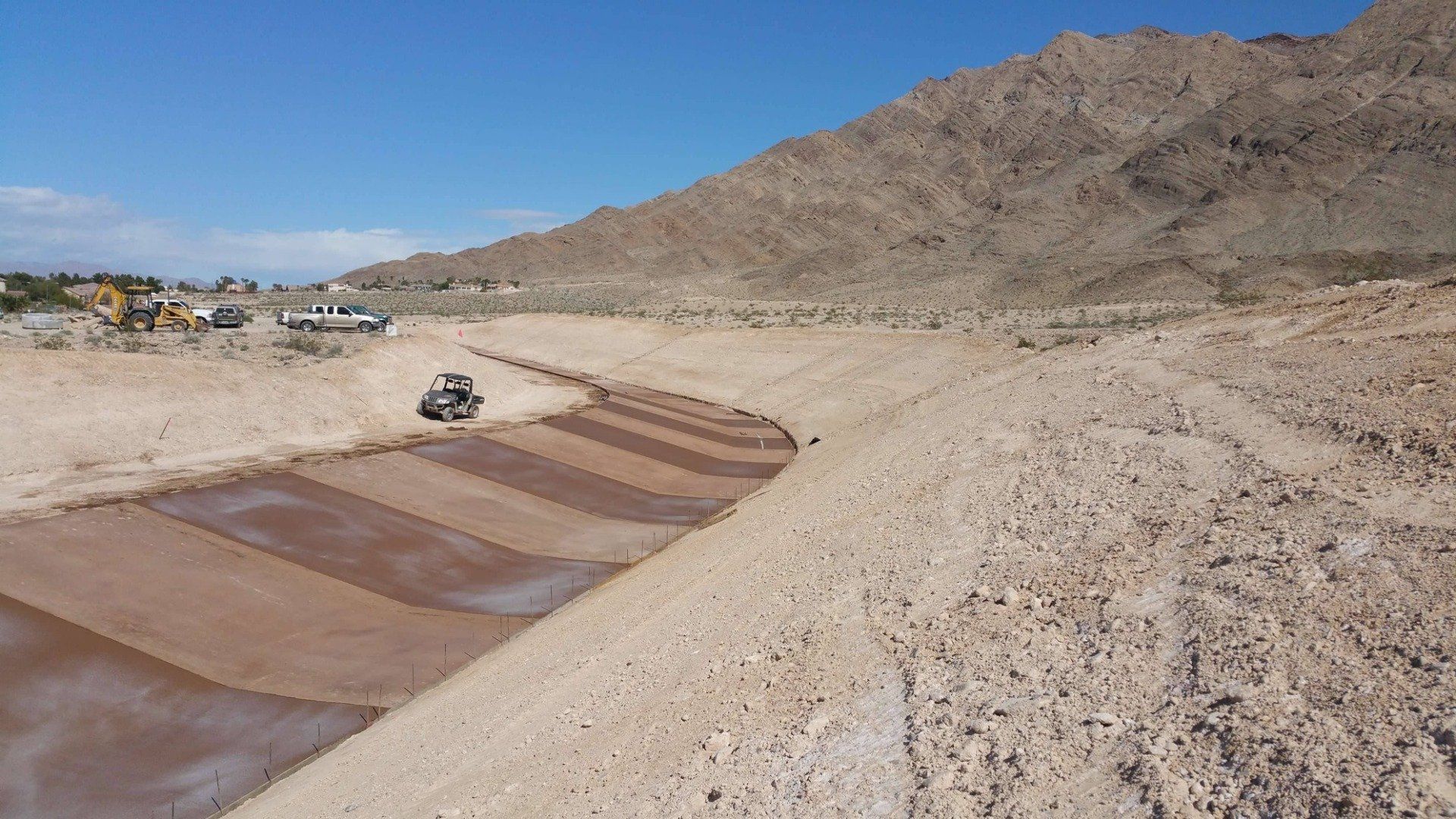 Tractor working in a large, earthen drainage channel near a mountain under a blue sky.
