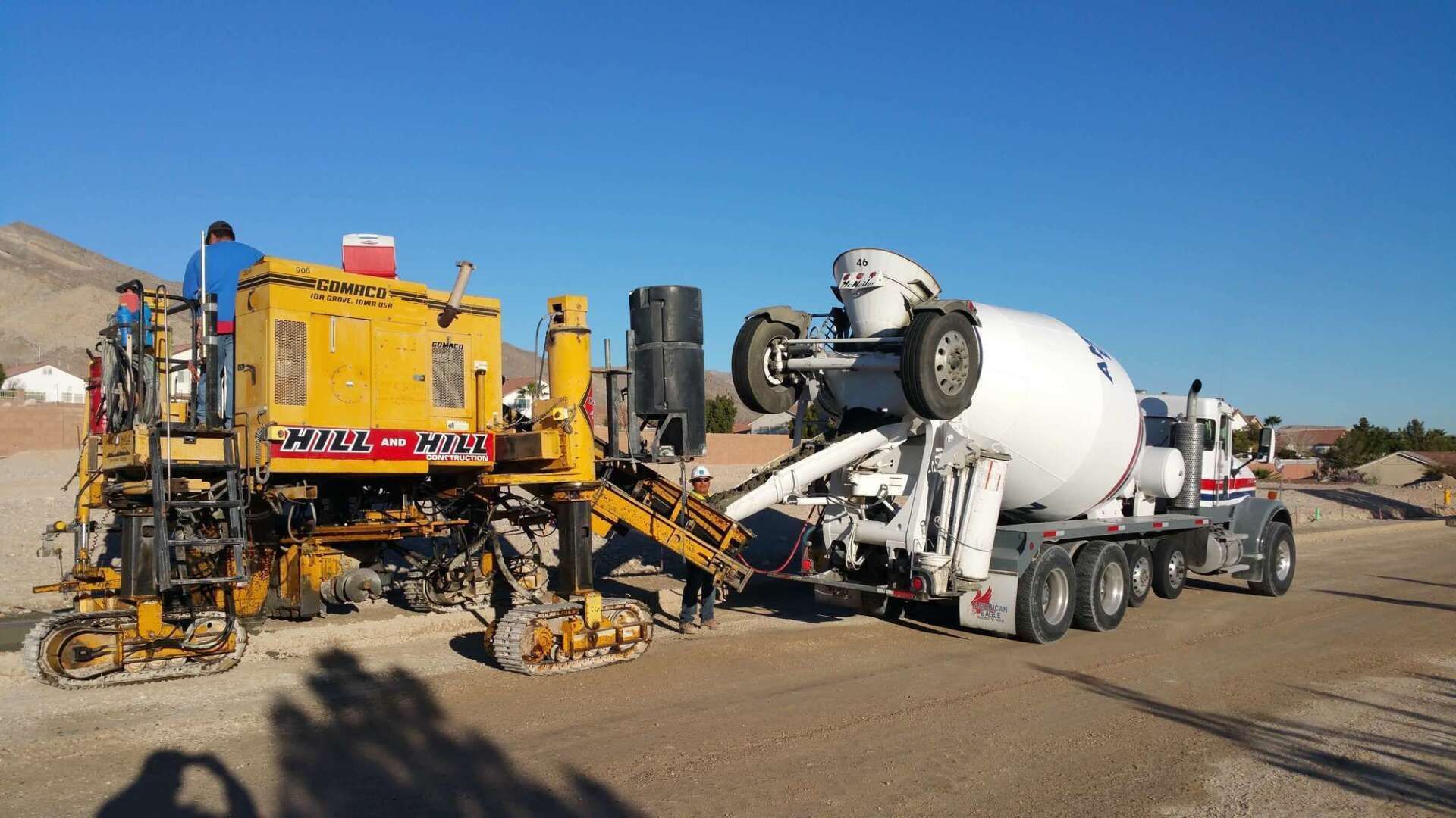 Concrete paving machinery laying road surface; truck pouring concrete. Outdoors, sunny.