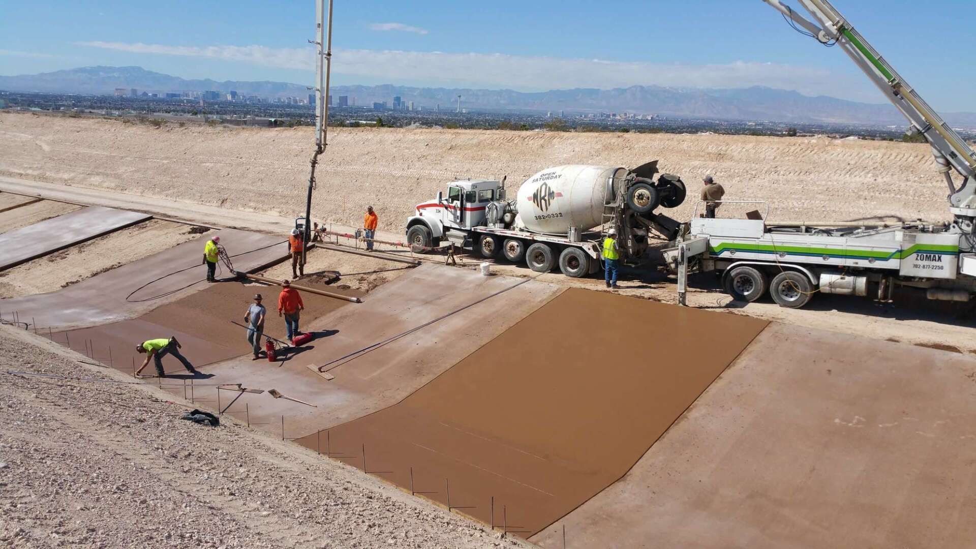 Construction workers pouring concrete at a site with a city skyline in the background. A cement truck and pump are present.