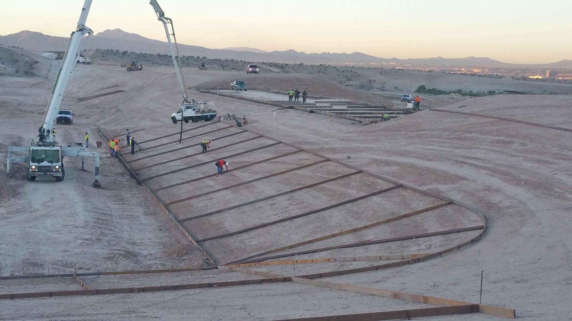Construction site with workers pouring concrete. Truck with boom, flat land, and prepared formwork visible.