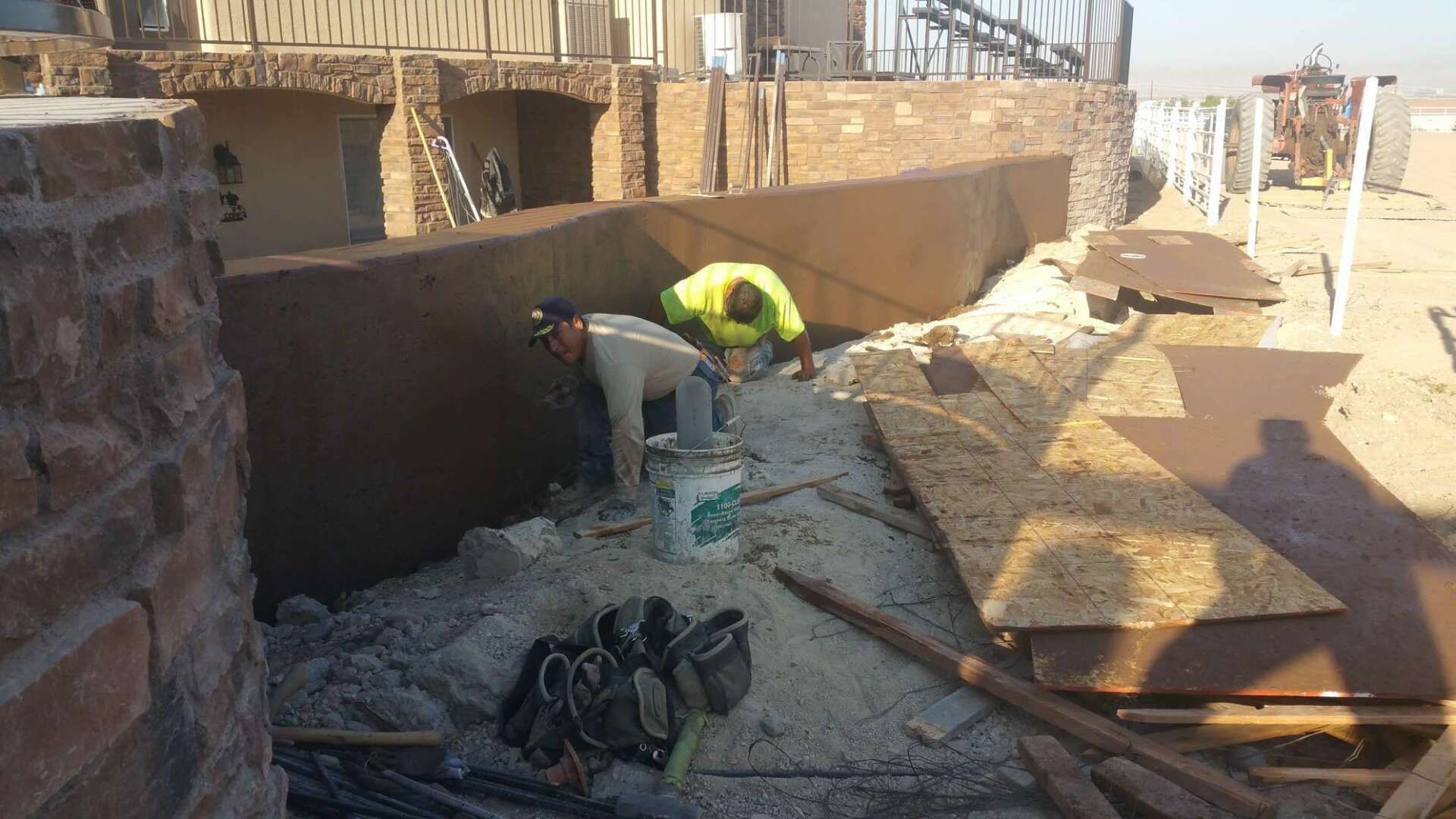 Two workers applying something to a large brown wall. Construction site with materials and tools.
