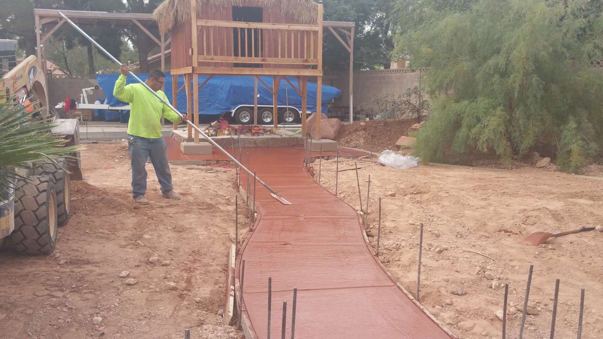 Man using a roller to smooth fresh, red-tinted concrete walkway. Construction site with a playhouse in the background.