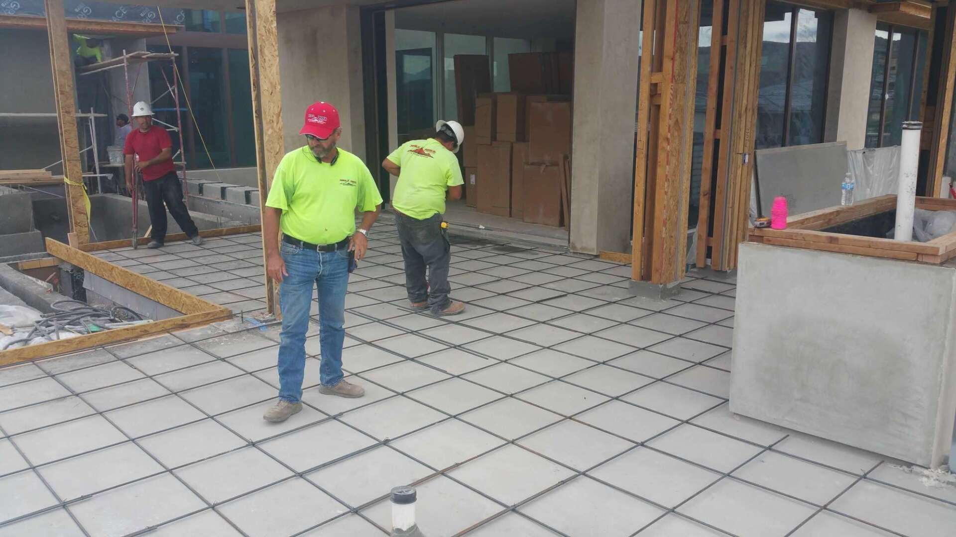 Construction workers on a concrete floor with rebar. Two in neon green shirts, one in red, working near a building's framing.