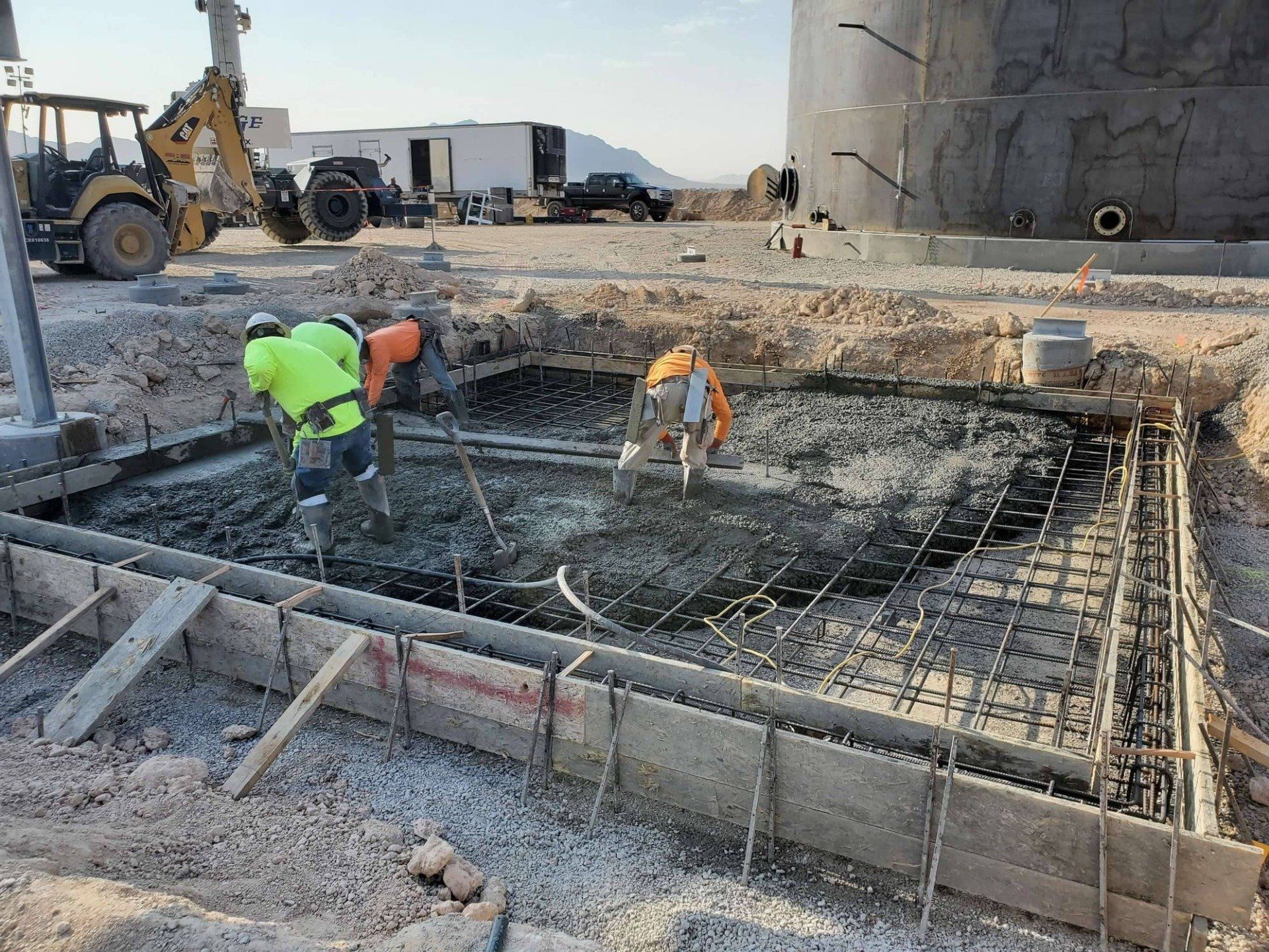 Construction workers pouring concrete into a rectangular form; a backhoe and storage tank are visible in the background.