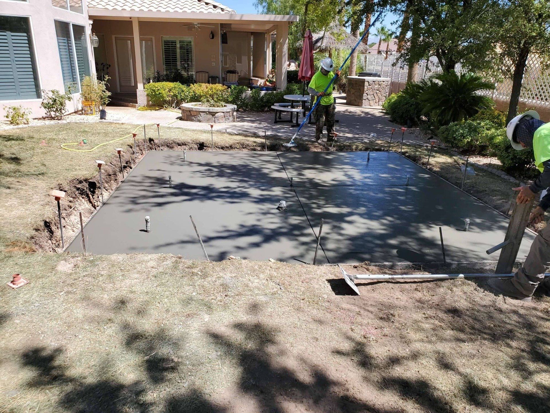 Workers pouring and leveling concrete slab in a backyard, near a house with a flag, on a sunny day.