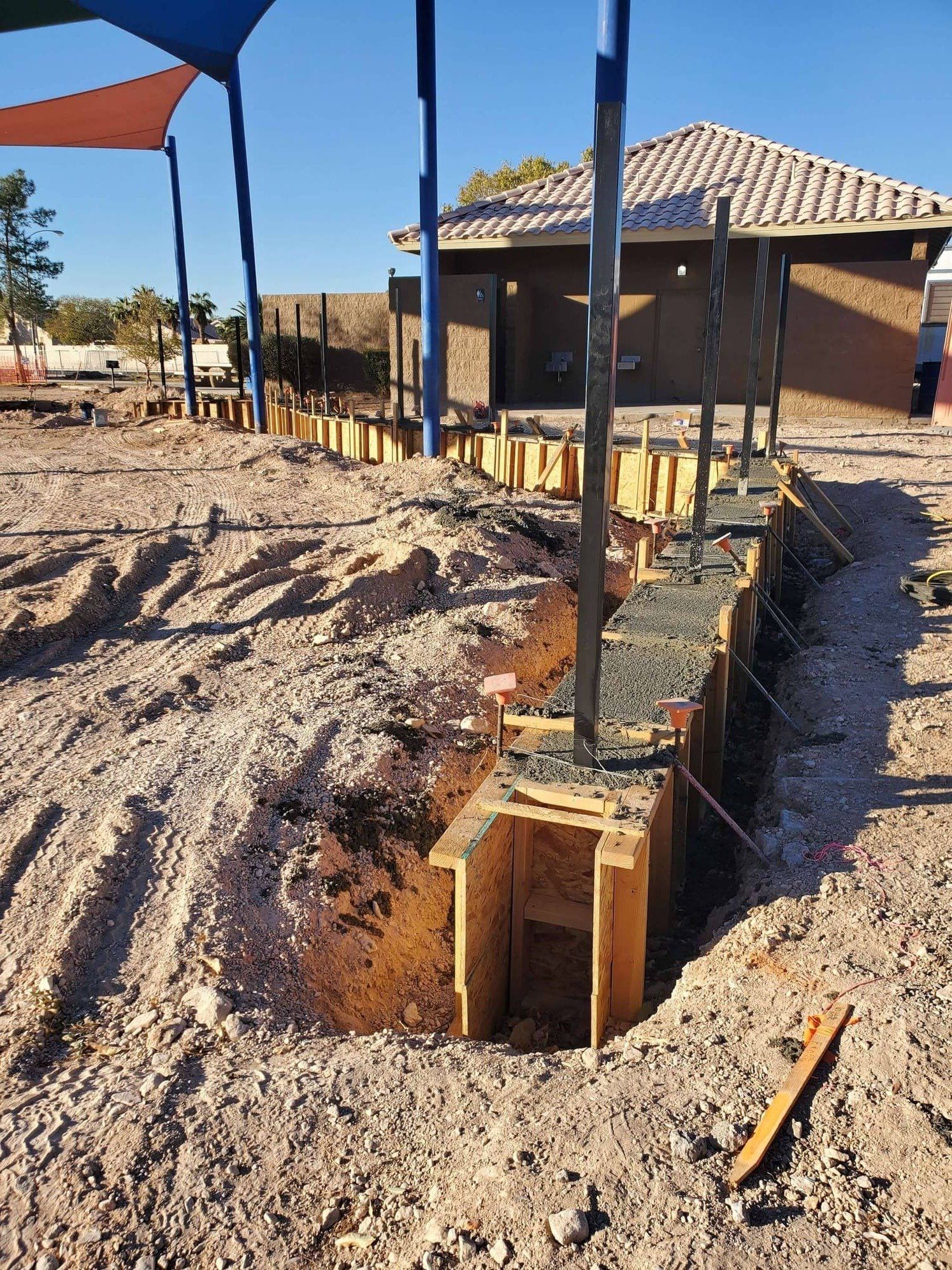 Construction site: wooden forms for a concrete foundation in dirt. A building with a tiled roof is in the background.