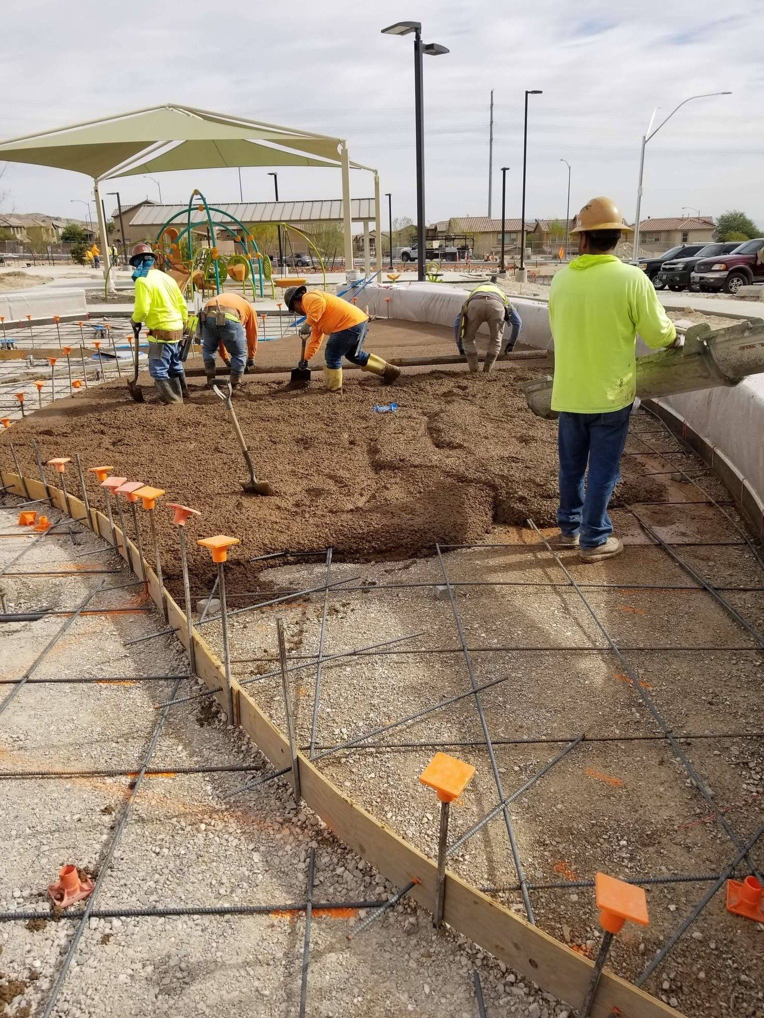 Construction workers pouring concrete at a park.  They're using shovels and a wheelbarrow.  Rebar and forms are visible.
