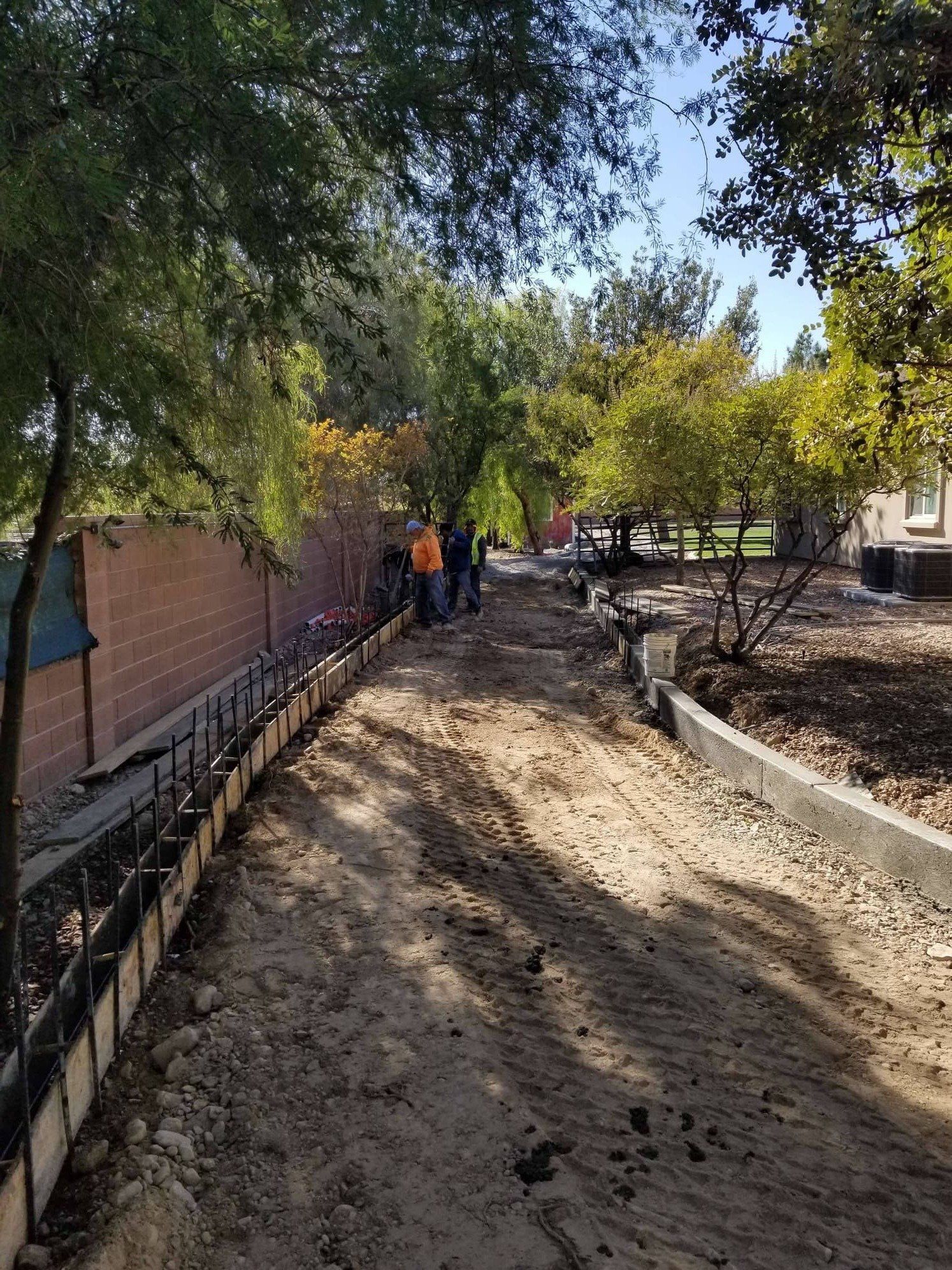 Construction of a pathway with workers, forms, and concrete along a brick wall and trees.