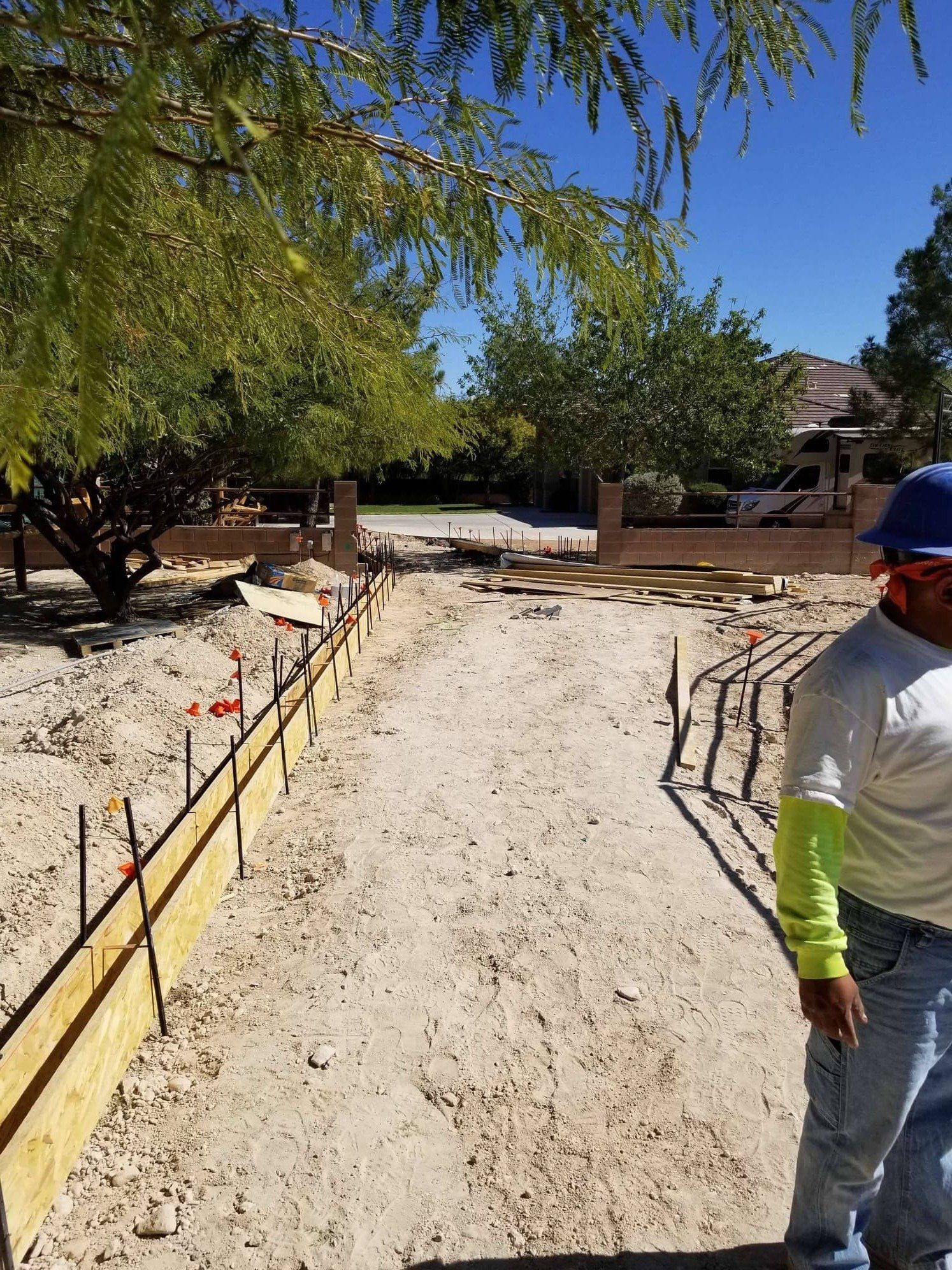 Construction site: Wooden forms outline a walkway. A worker in blue and jeans stands on the right. Sandy ground.