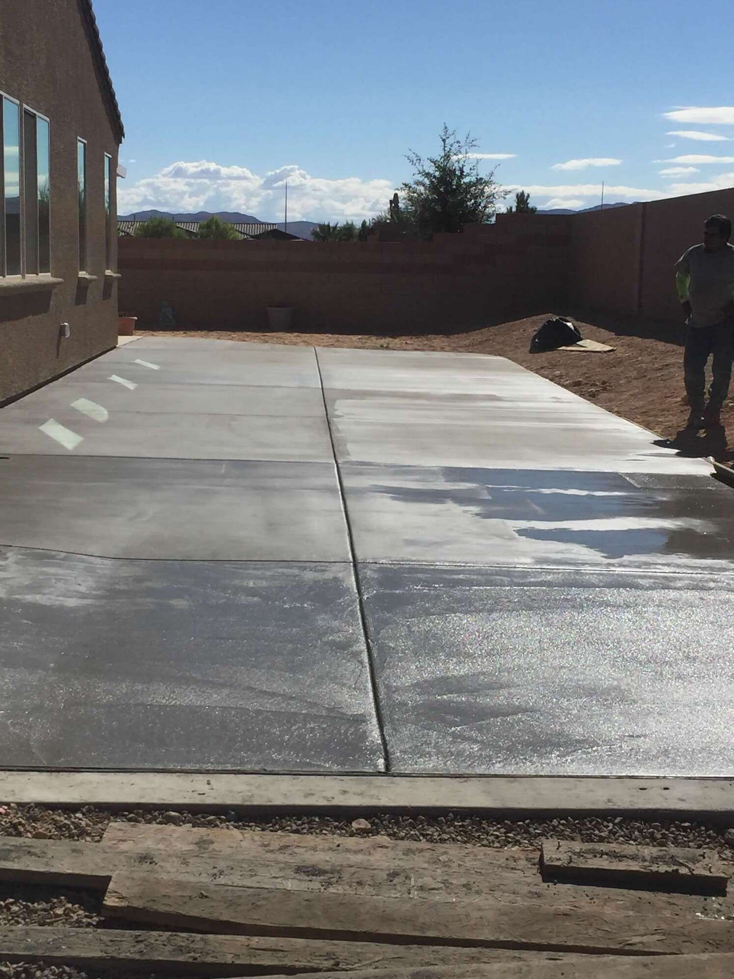 Backyard patio with fresh concrete; a man stands nearby under a blue sky.