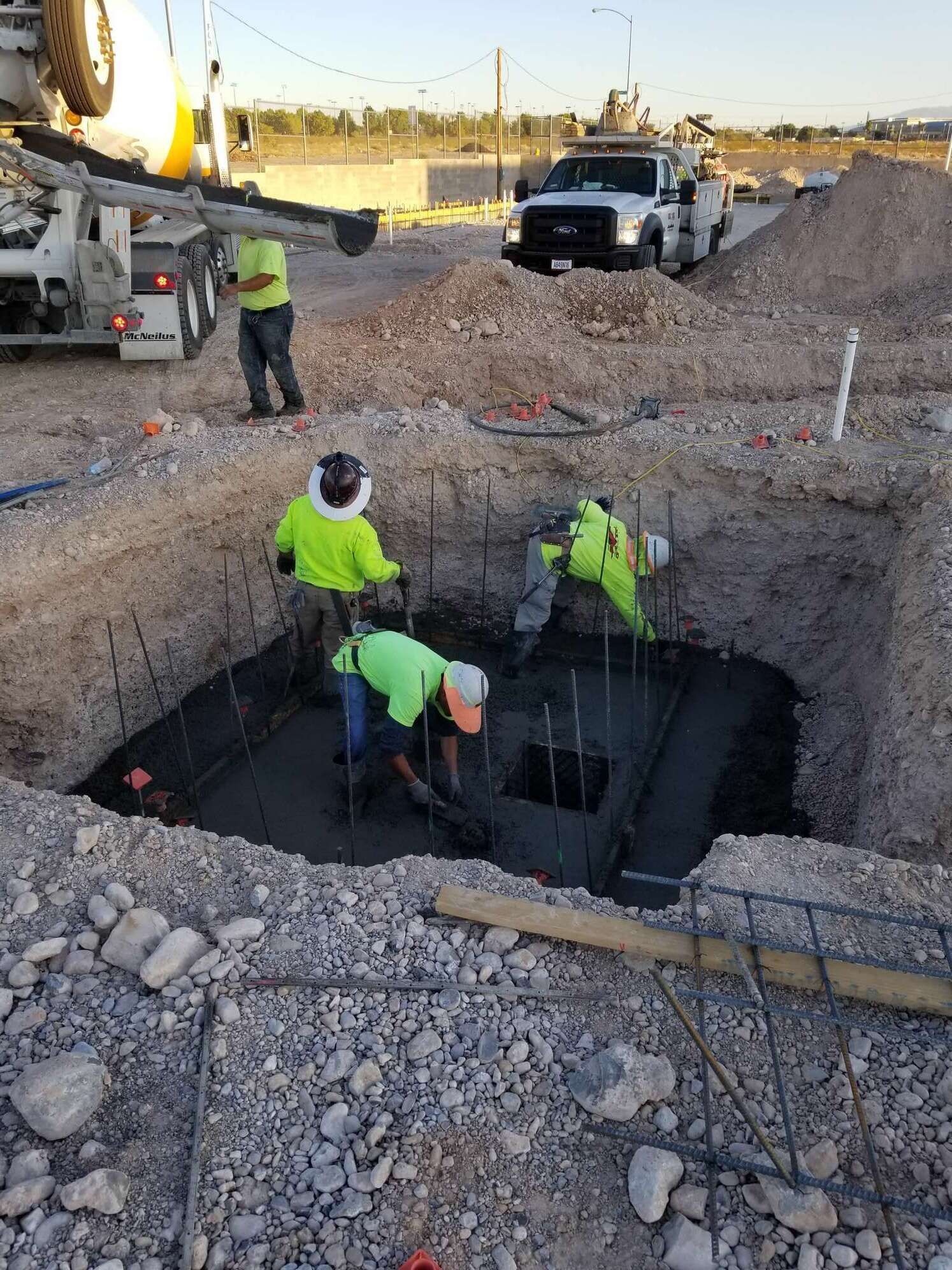 Construction workers pouring concrete into a foundation pit with rebar, next to a cement truck on a construction site.