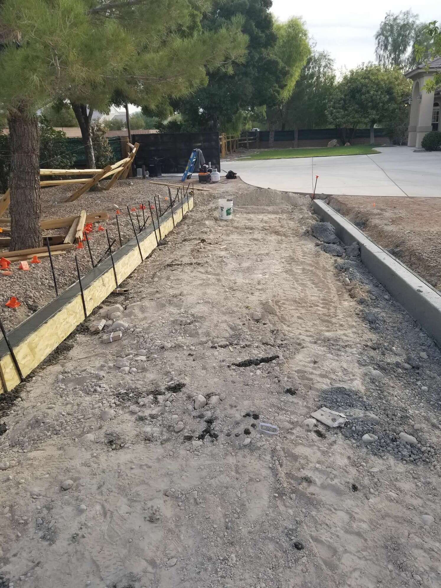 Gravel pathway under construction, bordered by wooden forms and a concrete curb. A house and trees are in the background.
