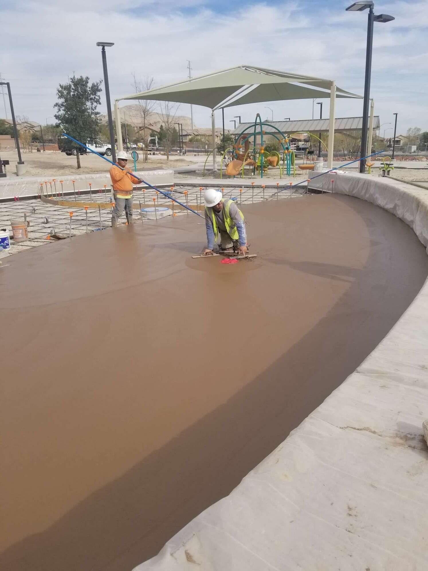 Construction workers smoothing wet concrete in a park area with a playground, under a shaded structure, on a sunny day.