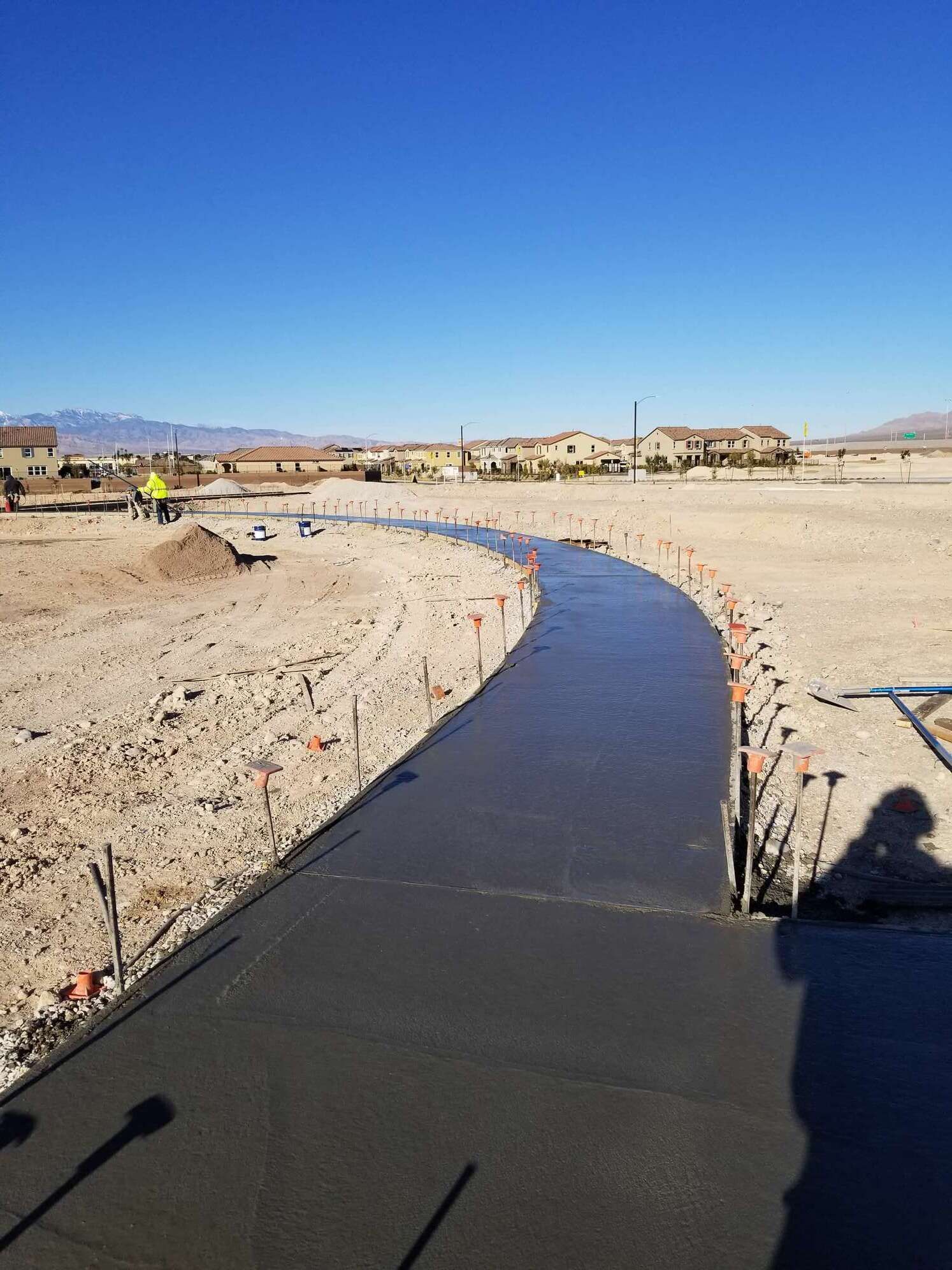 Newly poured concrete sidewalk curving through a dirt construction site under a clear blue sky.
