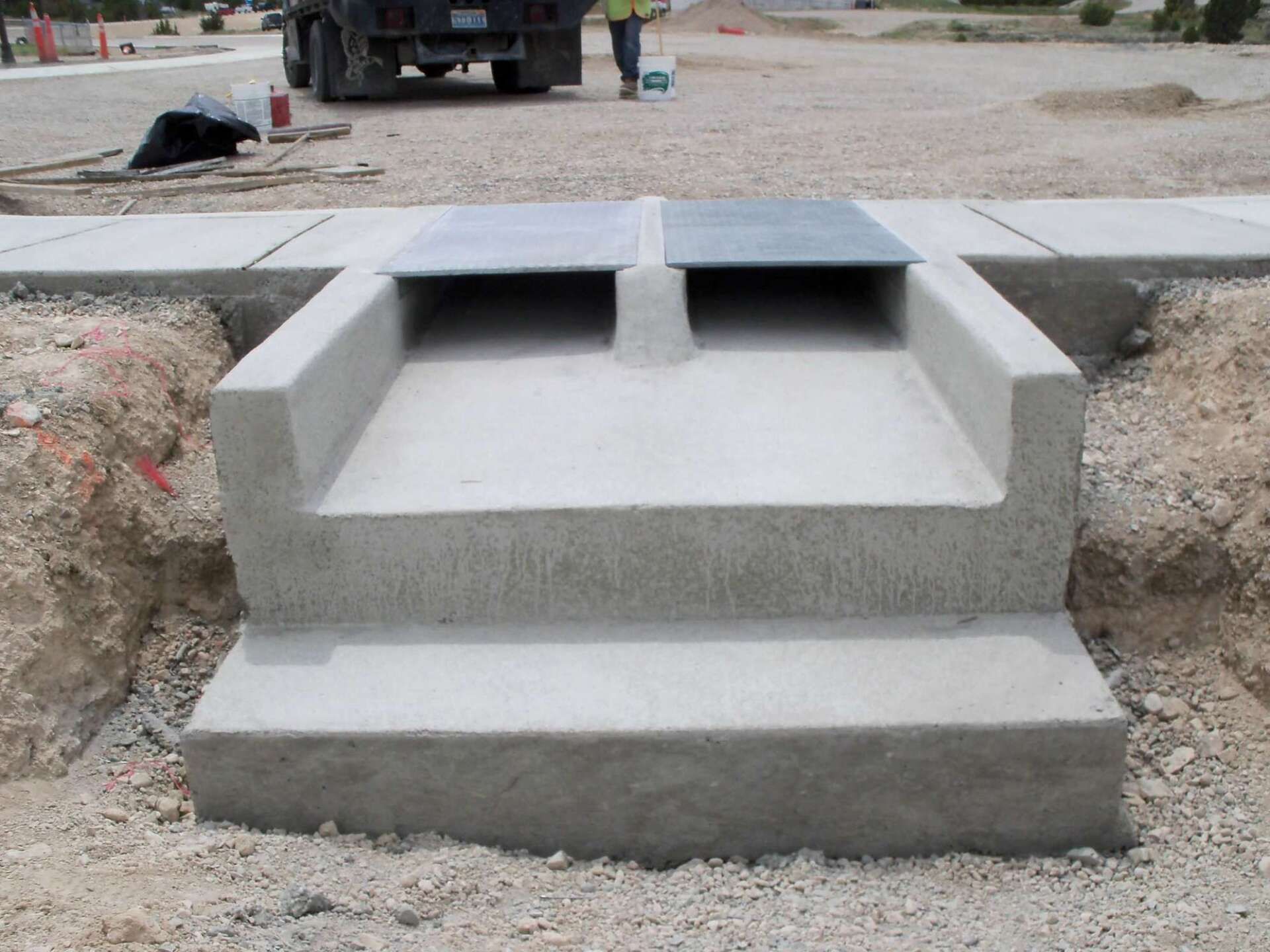 Concrete drainage structure with two openings, metal grate covers, and a truck in the background, construction site.