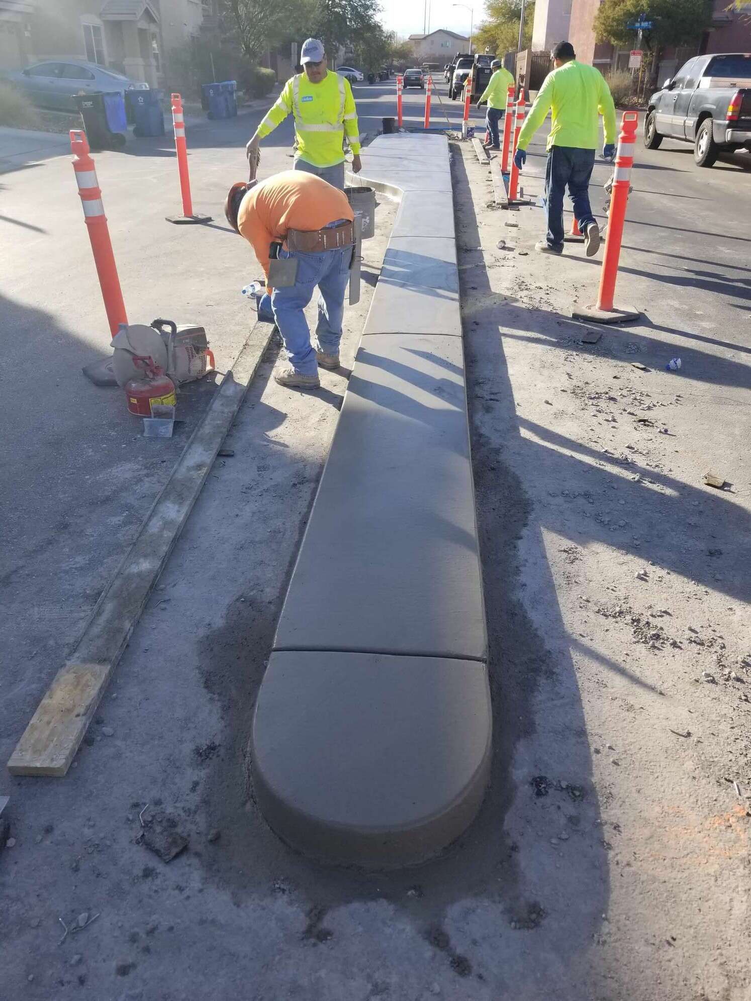 Construction workers pouring concrete curb on a residential street. Orange safety cones line the road.