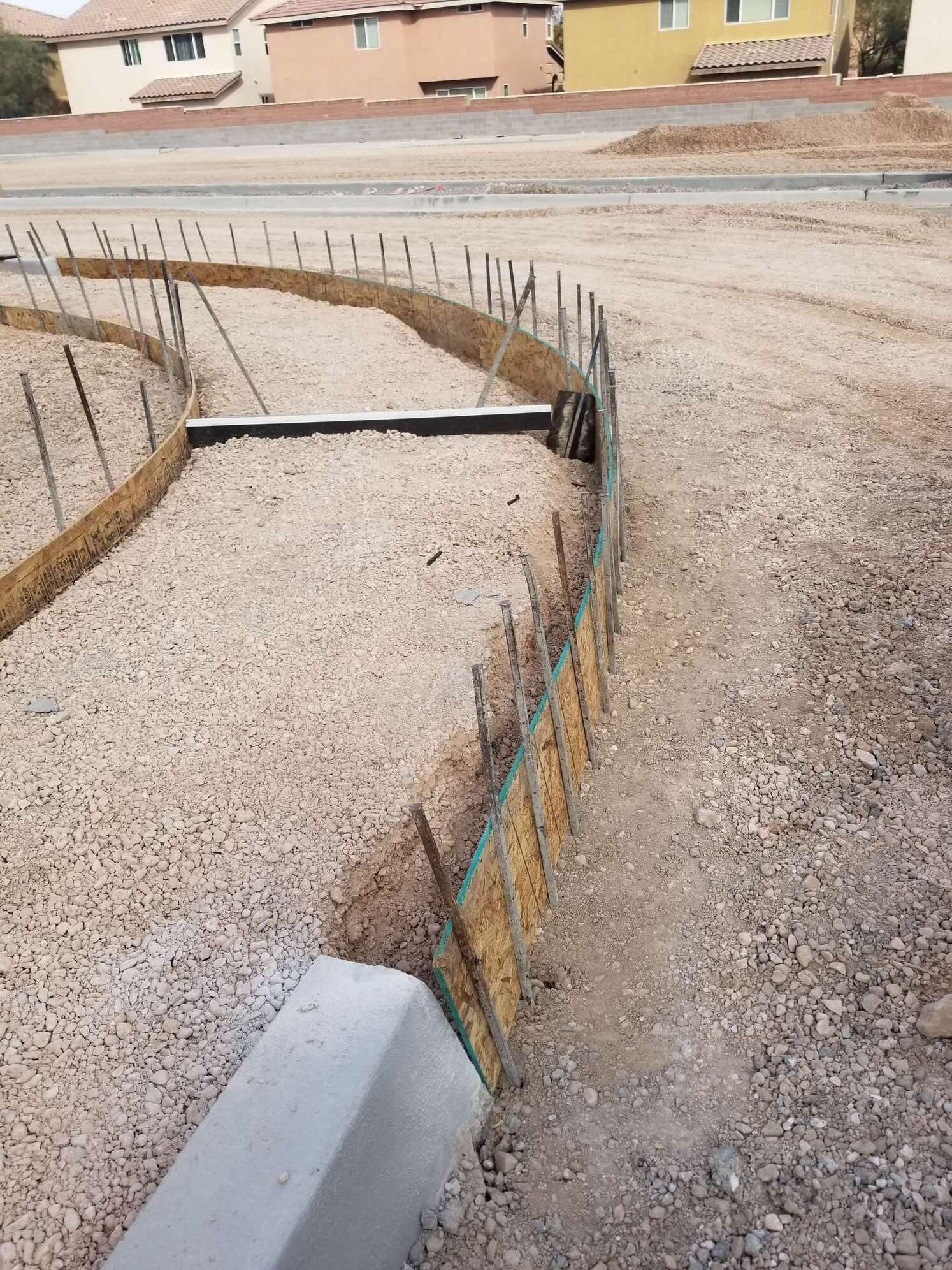 Construction site with gravel bed, border fence, concrete curb and houses in the background.