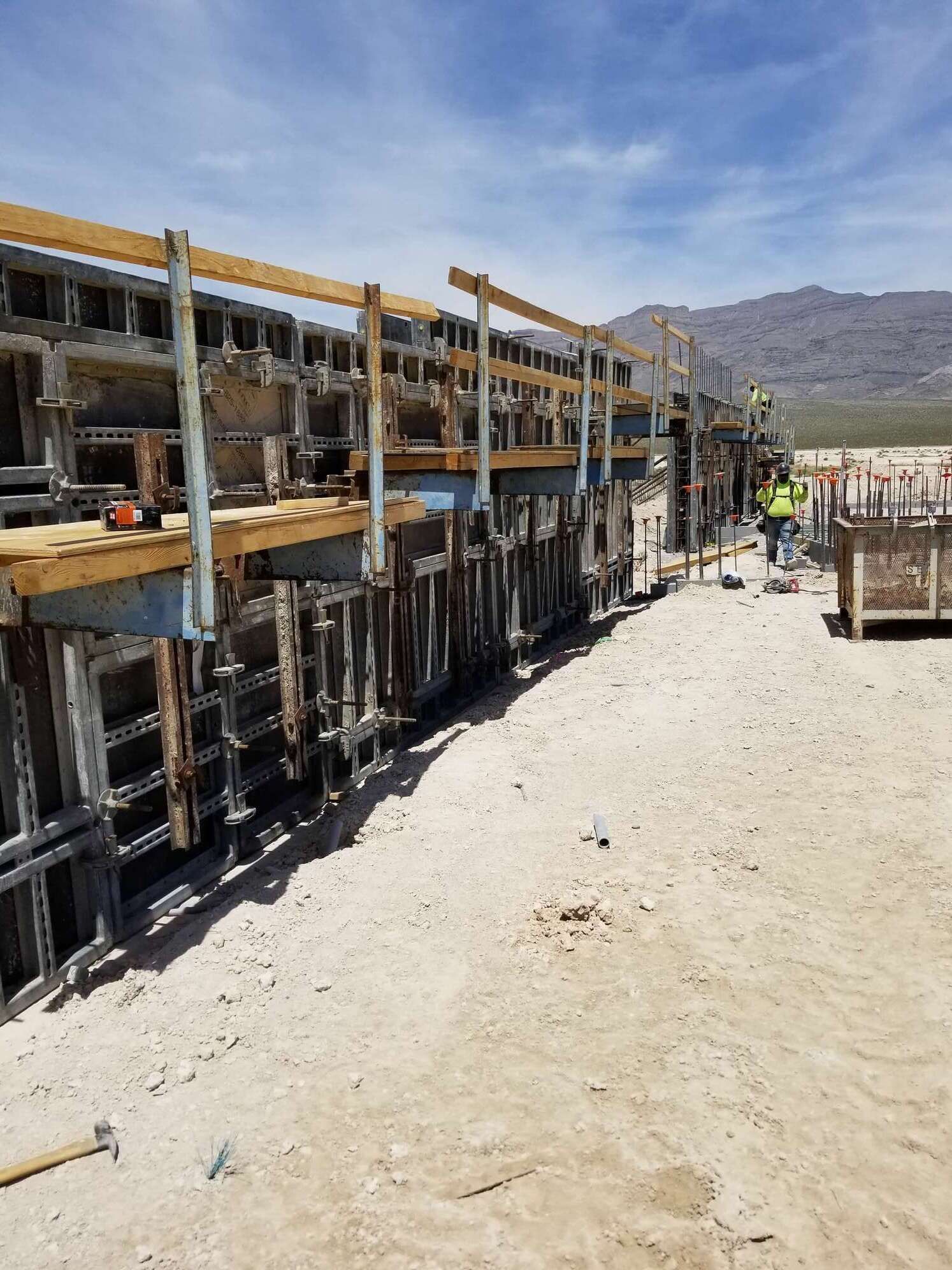 Construction site with concrete formwork, workers, and mountains in the background under a blue sky.