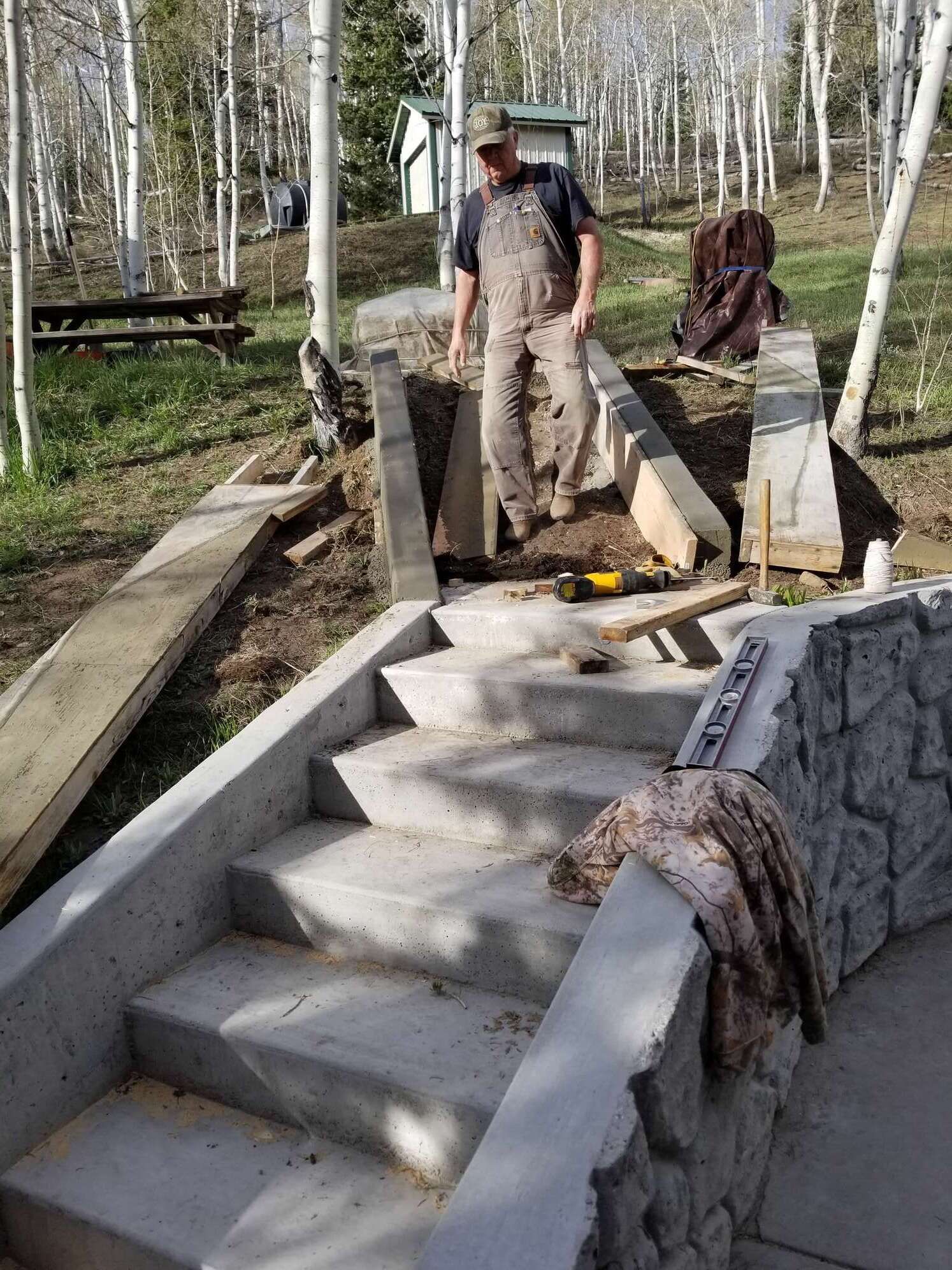 Man in overalls constructing concrete stairs with stone accents in a wooded area.