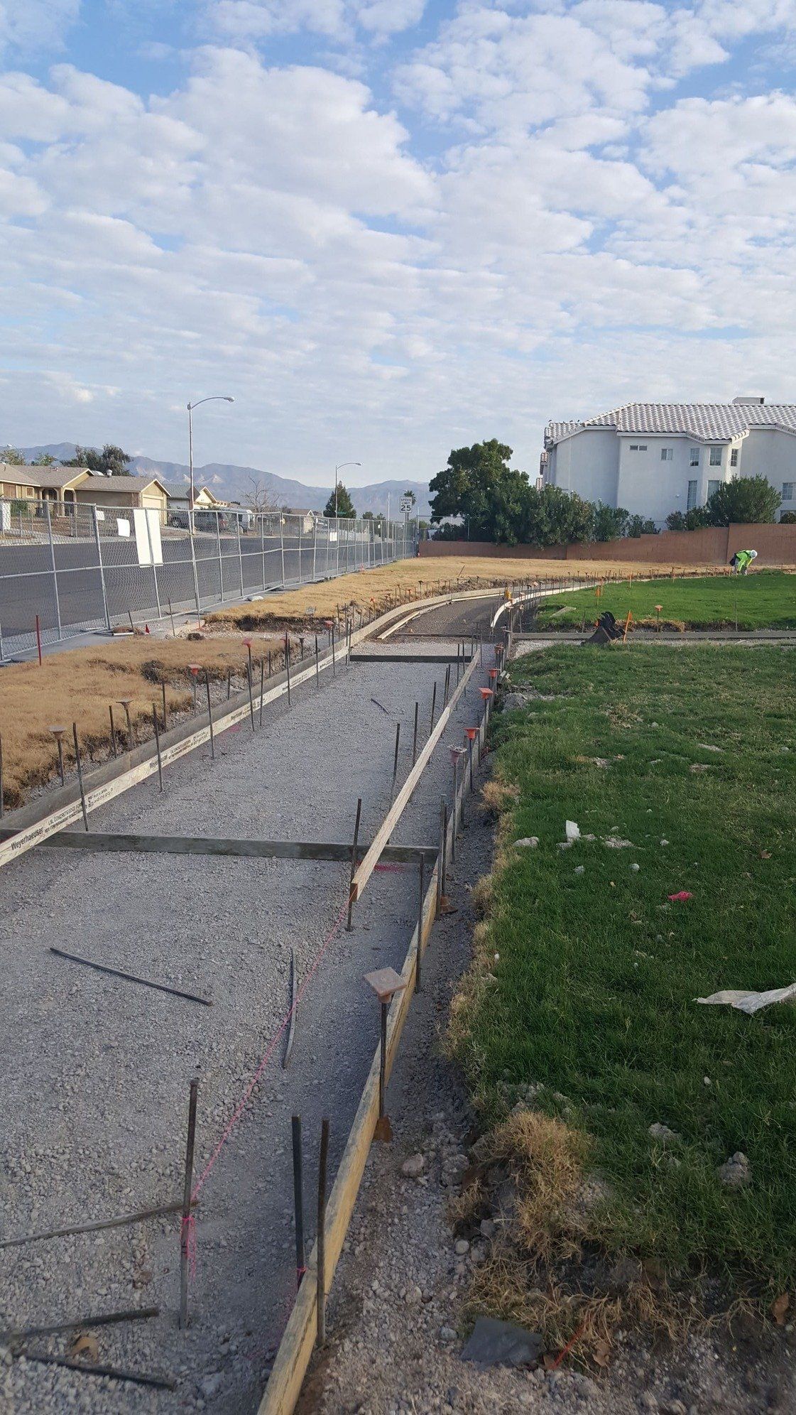 Concrete work site with gravel, wooden forms, and rebar next to green grass and a road.