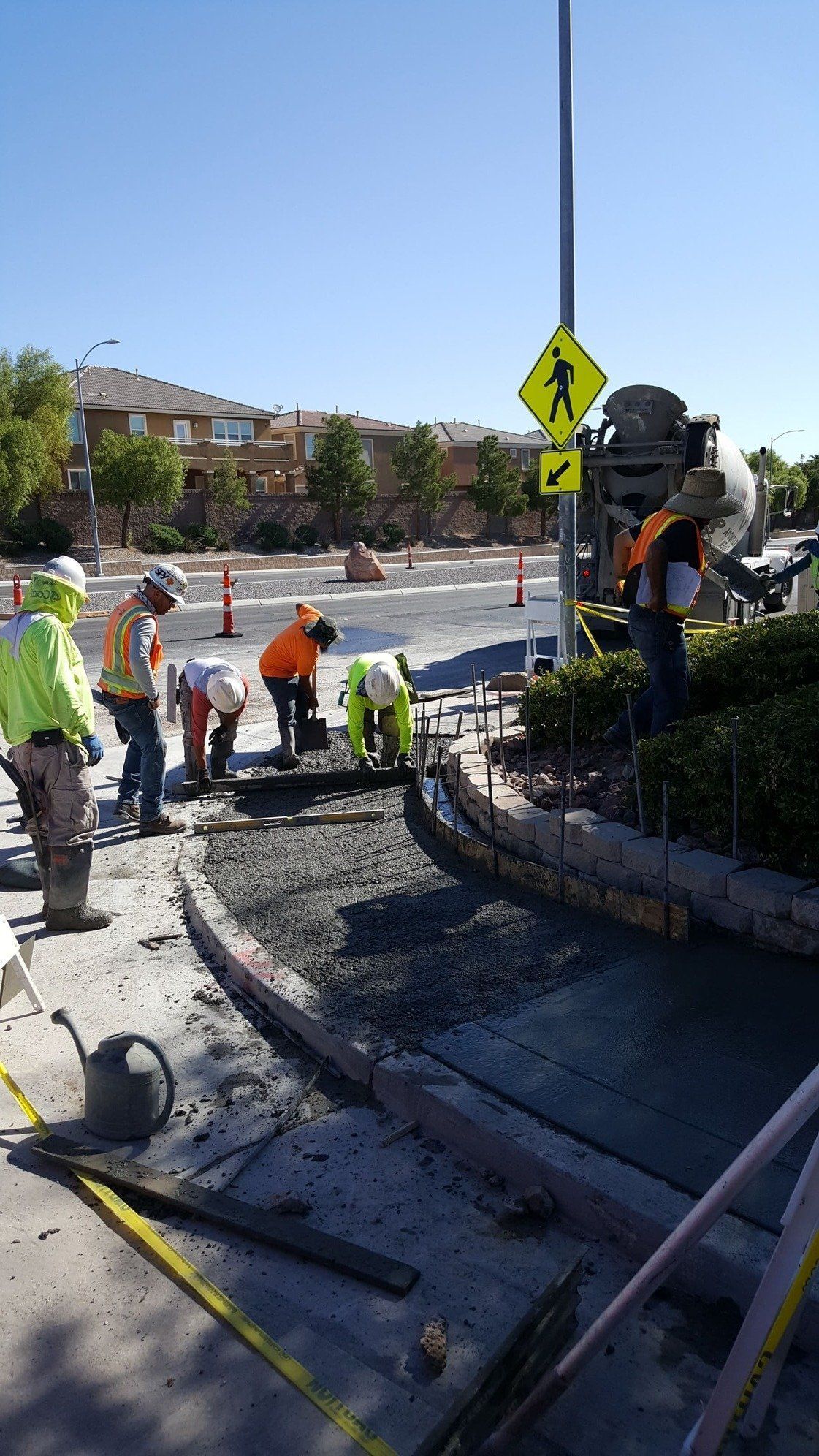 Construction workers pouring concrete on a sidewalk next to a road, with a crosswalk sign in the background.
