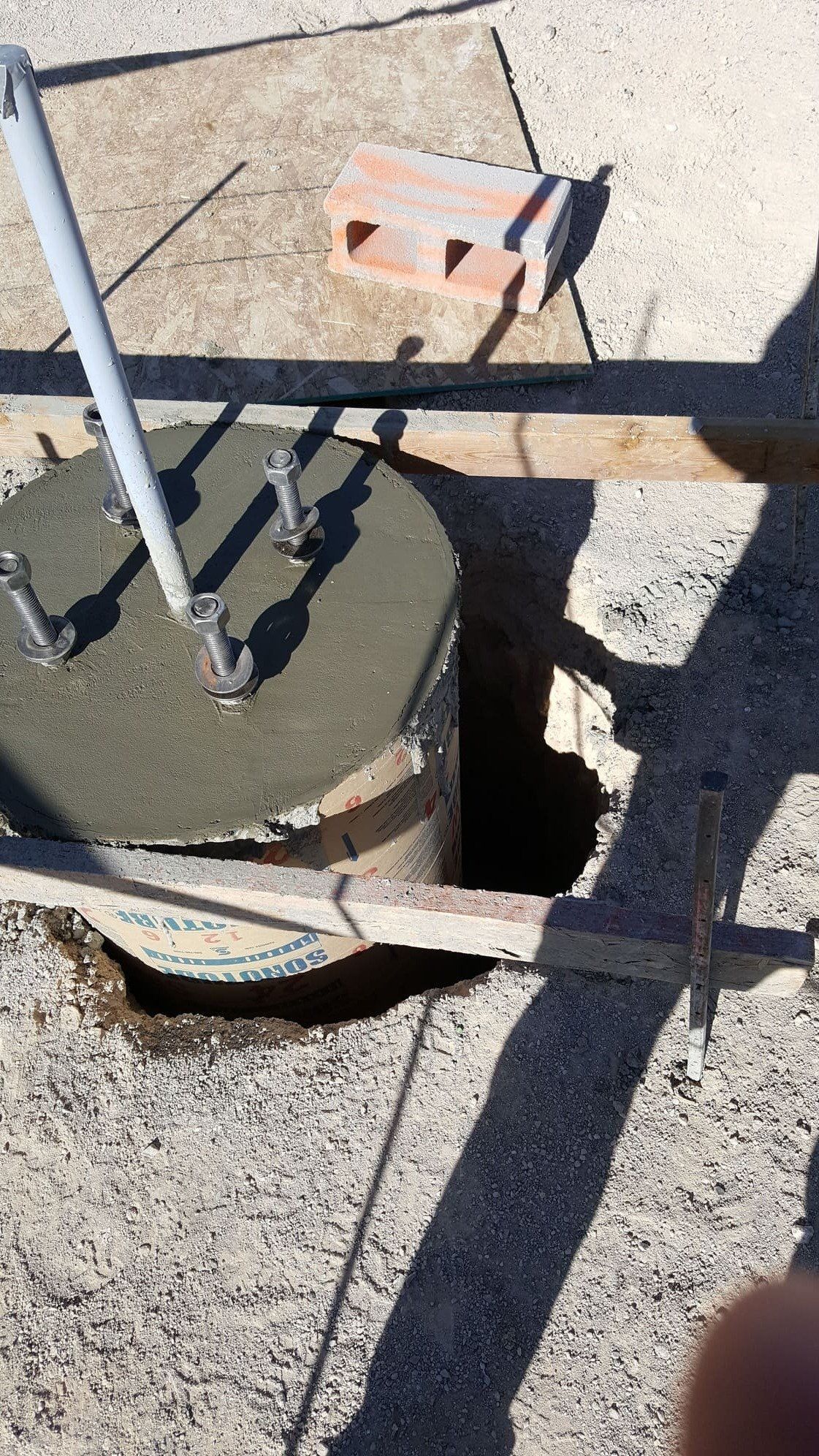 Concrete poured around bolts in a circular formwork, with a pole, bricks, and gravel visible.