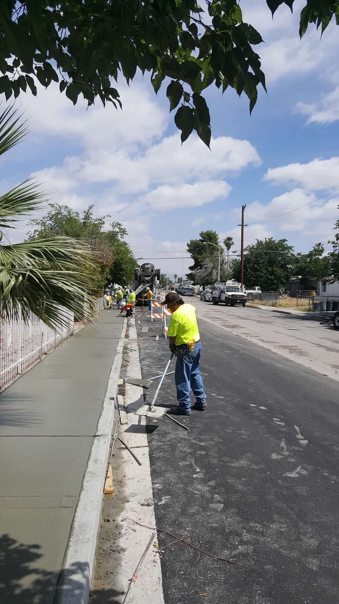 Construction workers paving a road and sidewalk on a sunny day. One worker in a yellow vest levels asphalt.