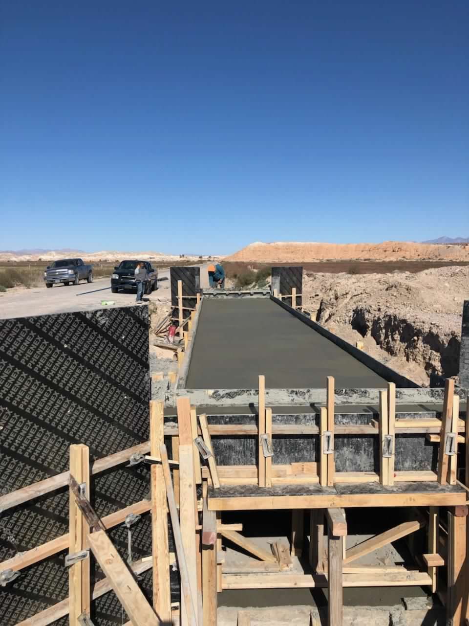 Concrete bridge construction site. Workers pour wet concrete into wooden forms near a road under a blue sky.
