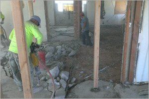 Workers demolishing a floor with jackhammers inside a building; wooden posts frame the view.