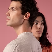 Un homme en chemise claire, regardant vers le haut, avec une femme derrière lui regardant légèrement sur le côté ; fond rose.