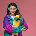 Femme tenant un skateboard jaune, portant un casque audio turquoise et une veste colorée, sur fond rose.