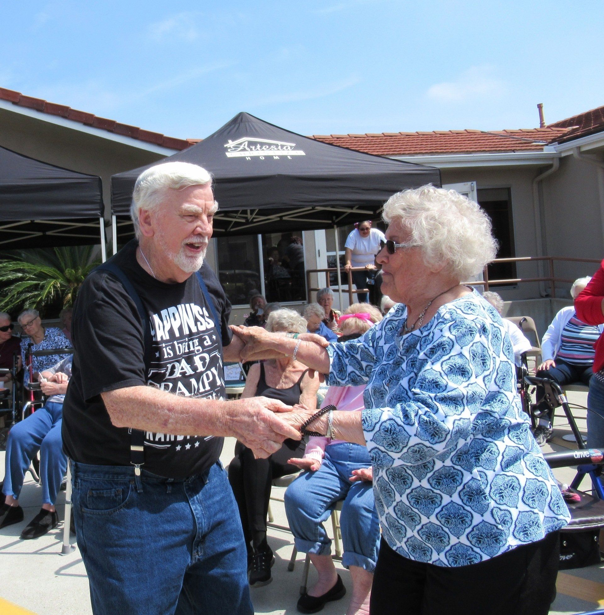 Elderly couple dancing outside at an event. Others watch from chairs.