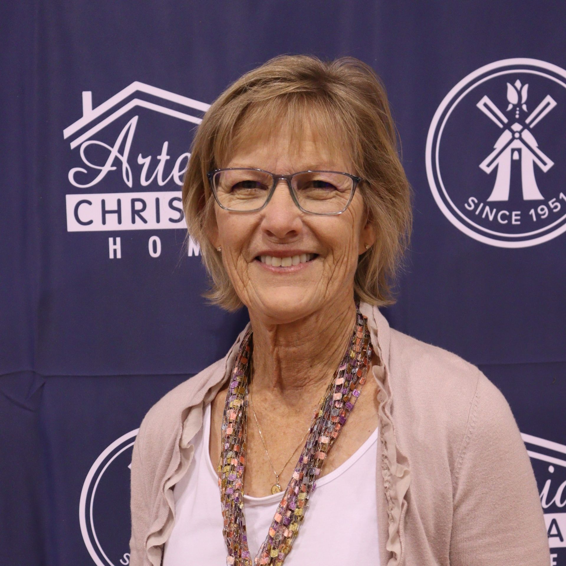 Woman with glasses smiling, wearing a cardigan and colorful scarf, in front of a logo-adorned backdrop.