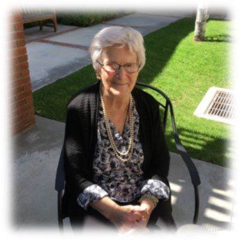 Elderly woman with white hair sits outside, wearing glasses and pearls.