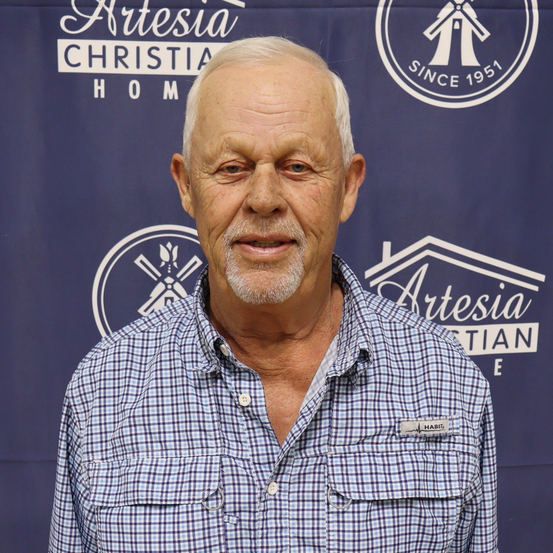 A smiling older man with gray hair and a beard, wearing a blue plaid shirt, in front of a banner for Artesia Christian Home.