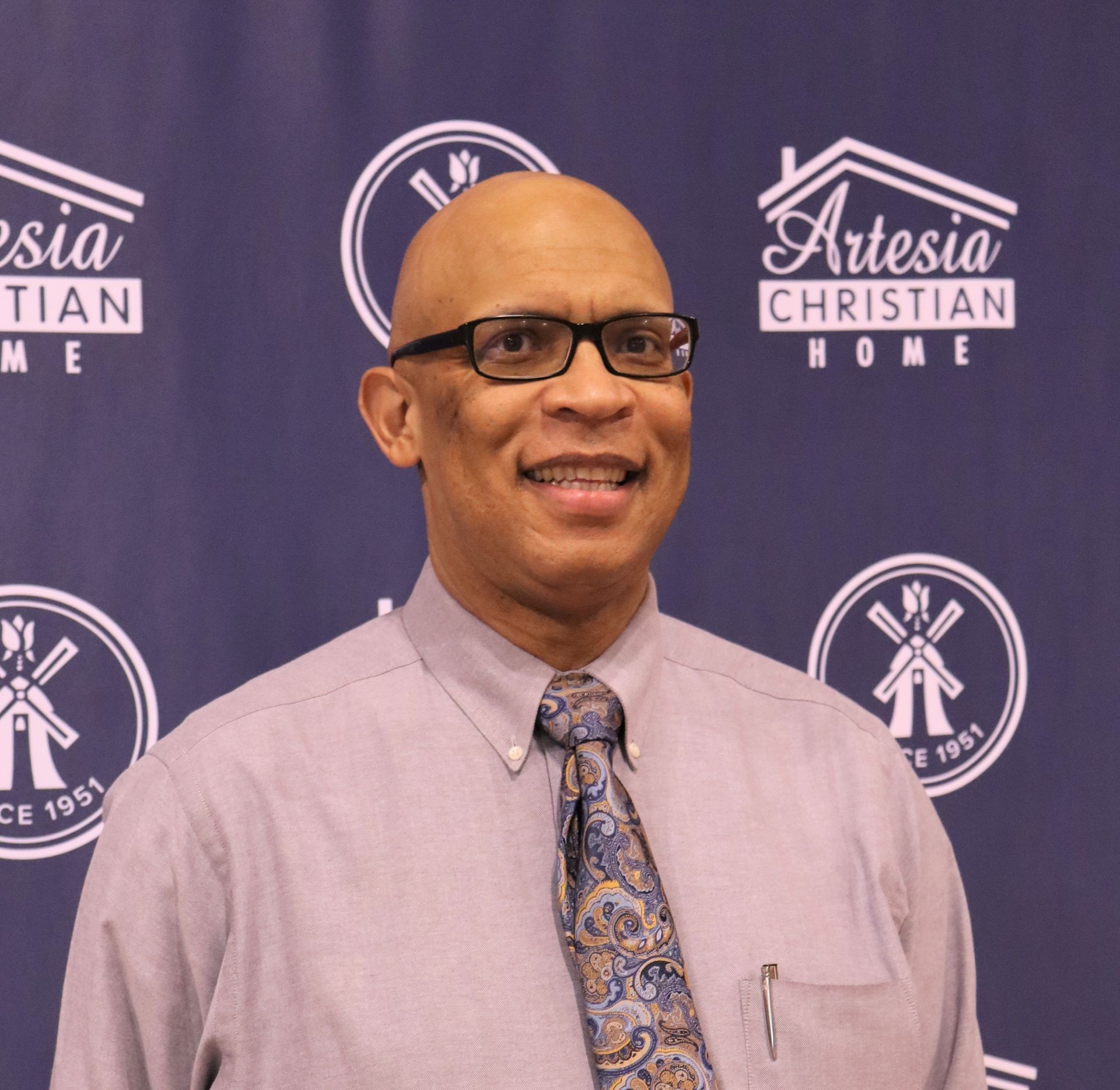 Bald man wearing glasses, smiling, wearing a button-down shirt and tie, in front of a banner with a logo.