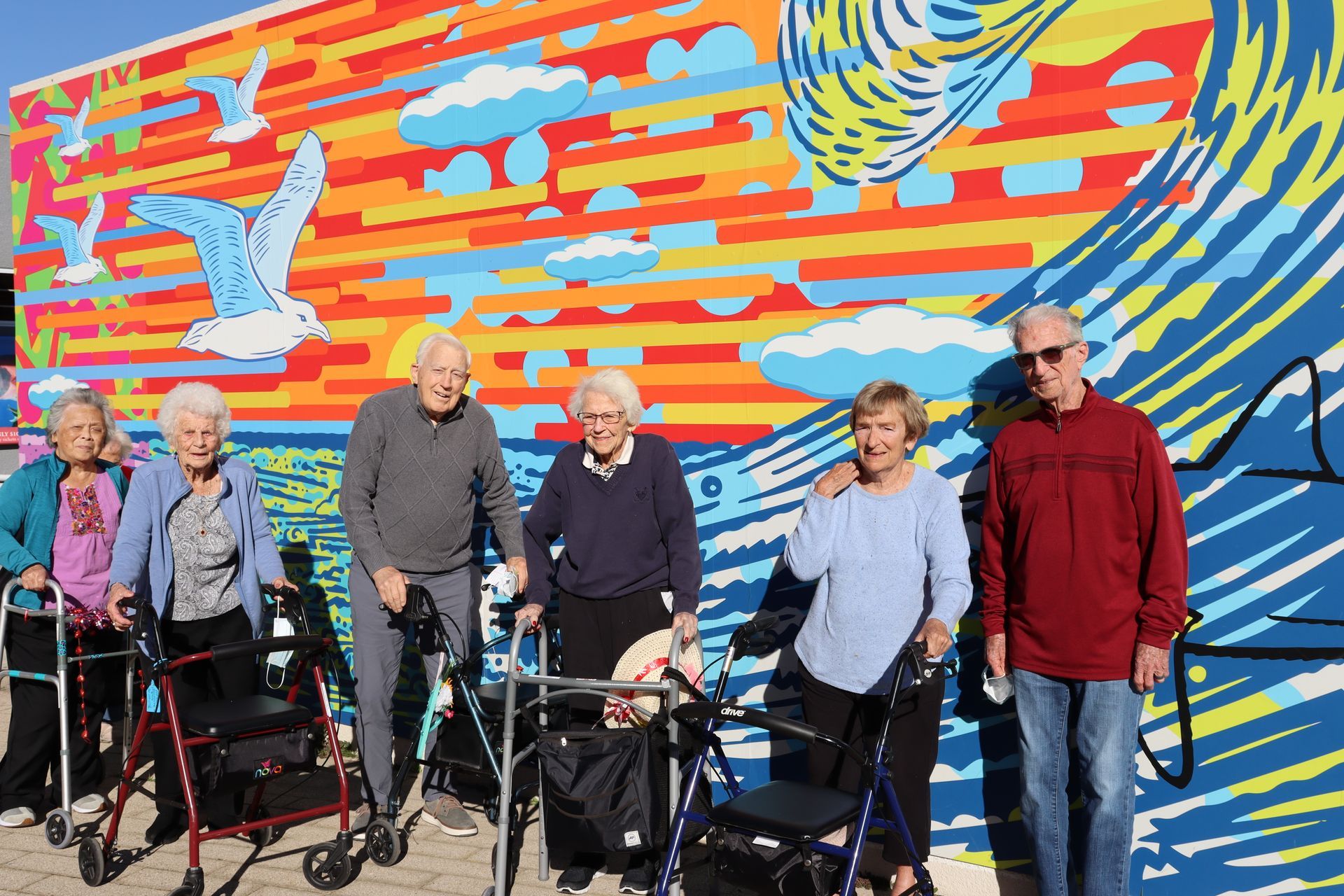 Group of seniors with walkers posing in front of a colorful mural of a beach scene.