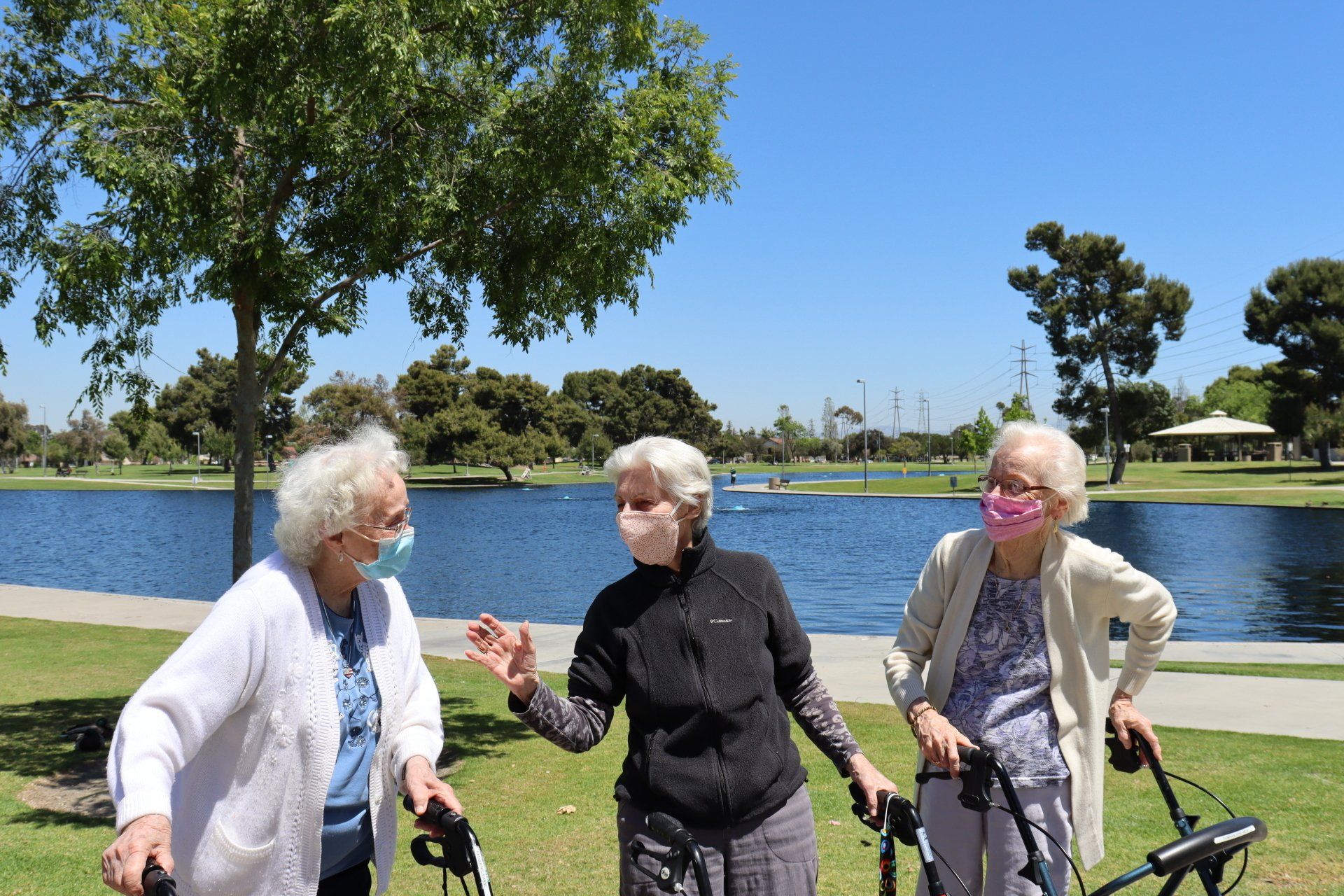 Three older women with walkers, masked, in a park by a lake, conversing, sunny day.