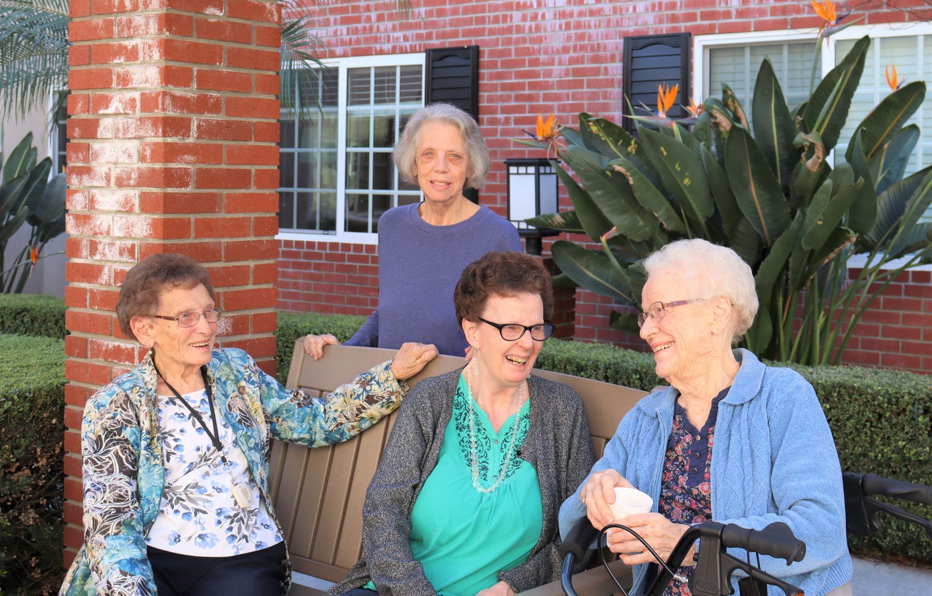 Four senior women laughing together outside, near a brick column and greenery.