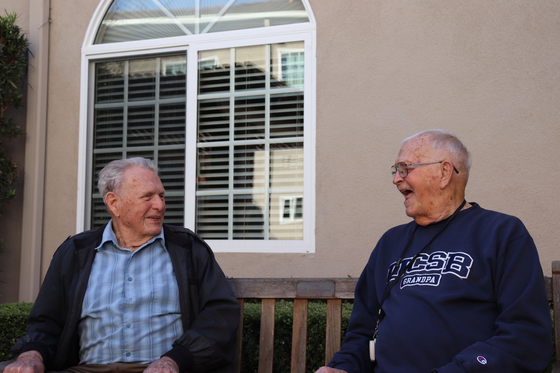 Two elderly men laugh, sitting on a bench outside. 