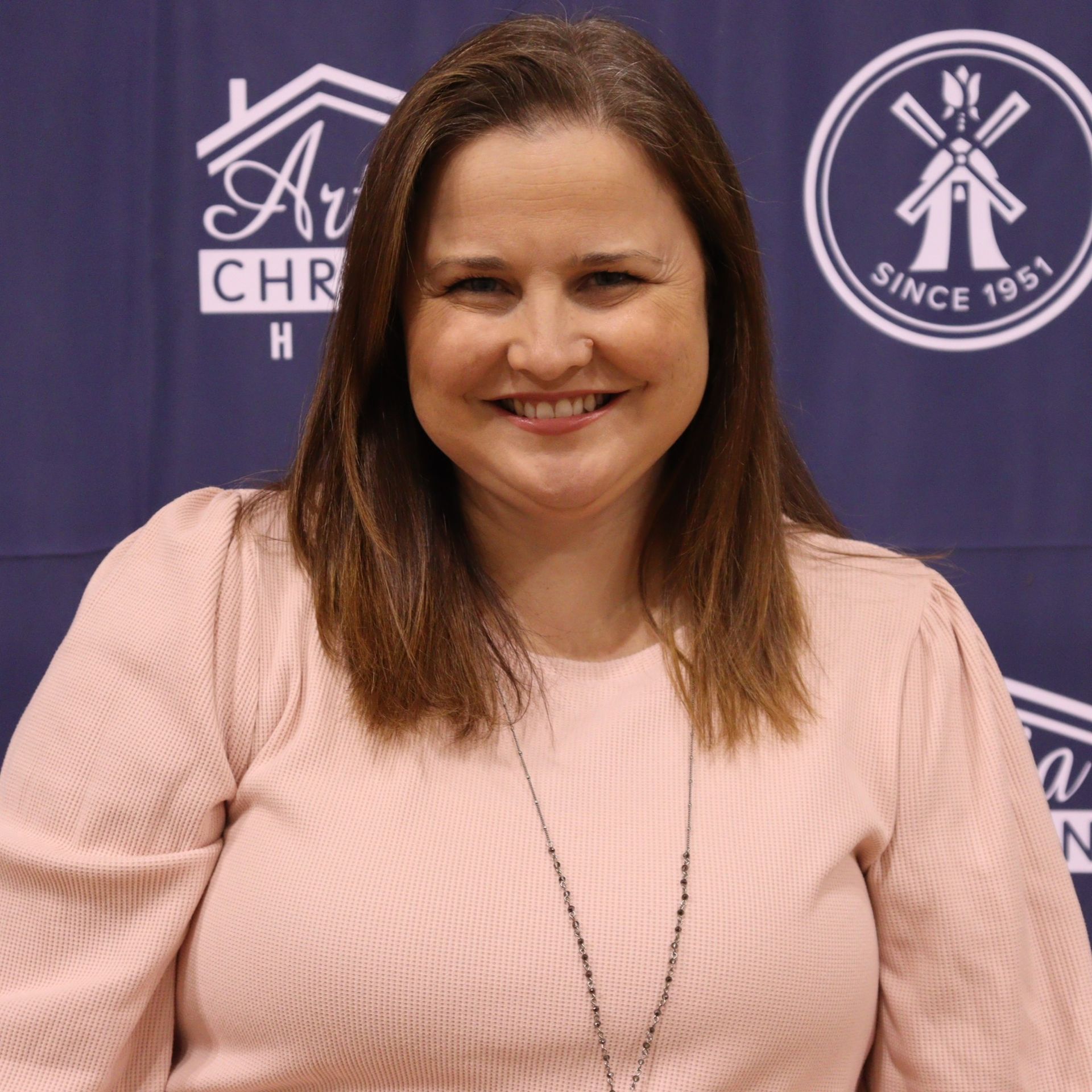 Woman with brown hair smiles, wearing a pink shirt and necklace, in front of a branded backdrop.
