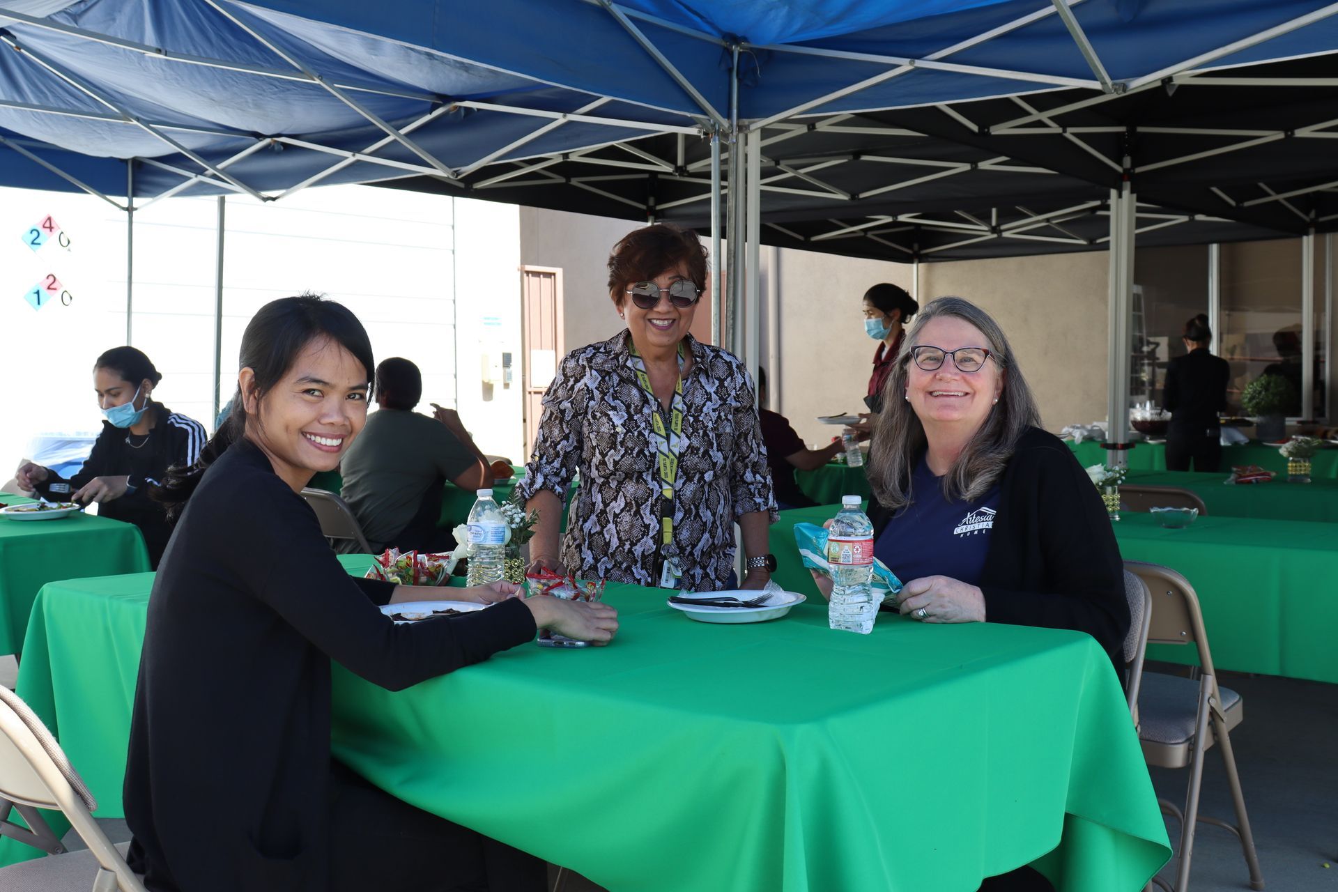 Three women smiling at a table covered in green cloth under a blue canopy.